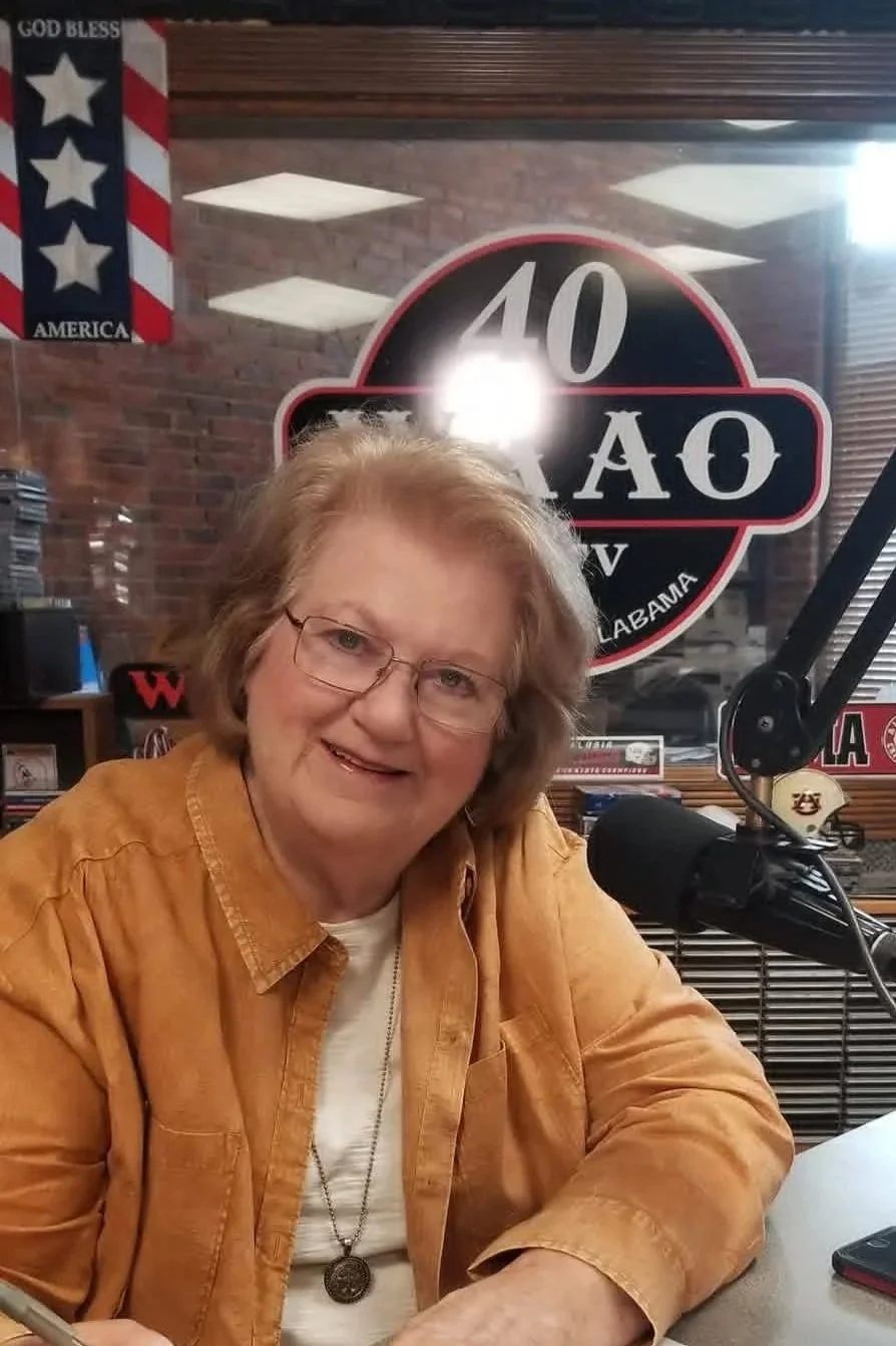 A woman with glasses and shoulder-length hair, wearing a brown jacket and a necklace, sitting in front of a microphone, smiling at the camera in a radio studio decorated with Alabama and patriotic signs.