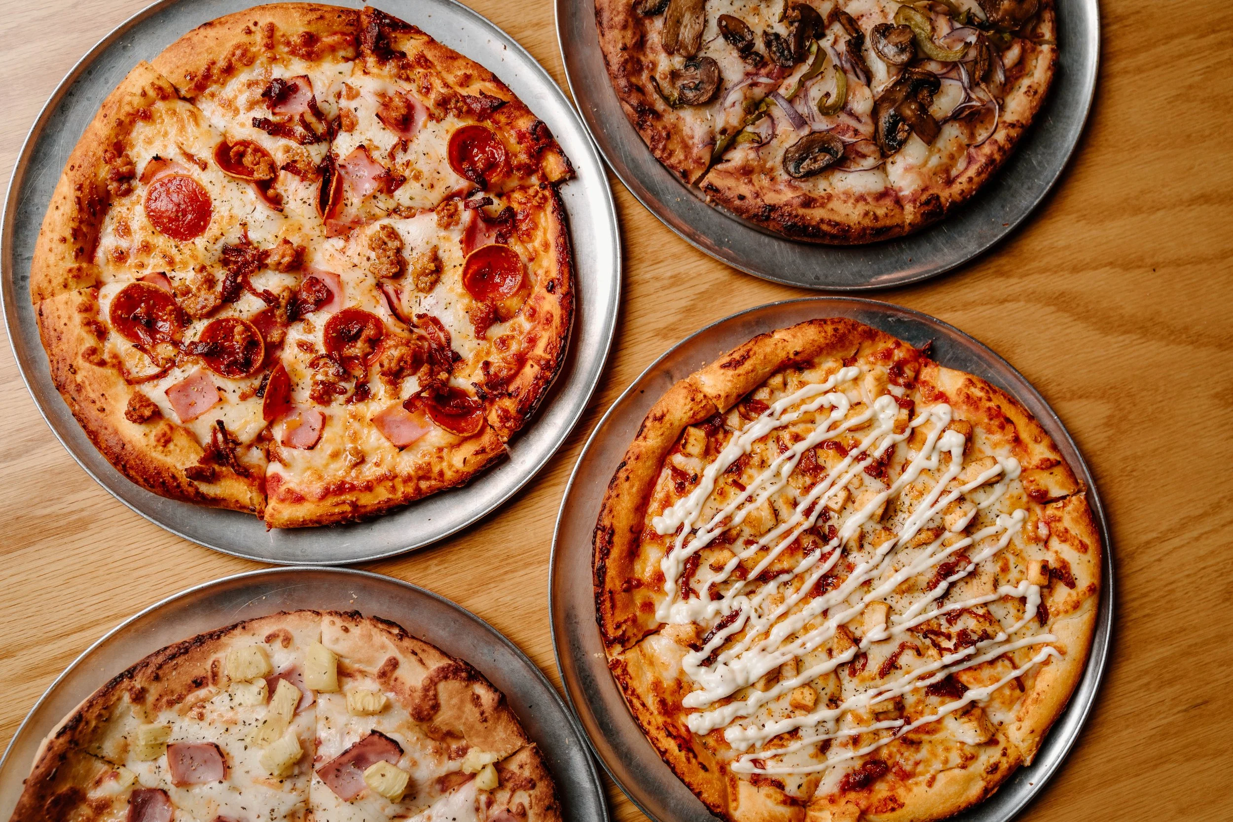Four pizzas on metal trays on a wooden table. The pizzas have various toppings including pepperoni, ham, mushrooms, pineapple, and drizzled white sauce.