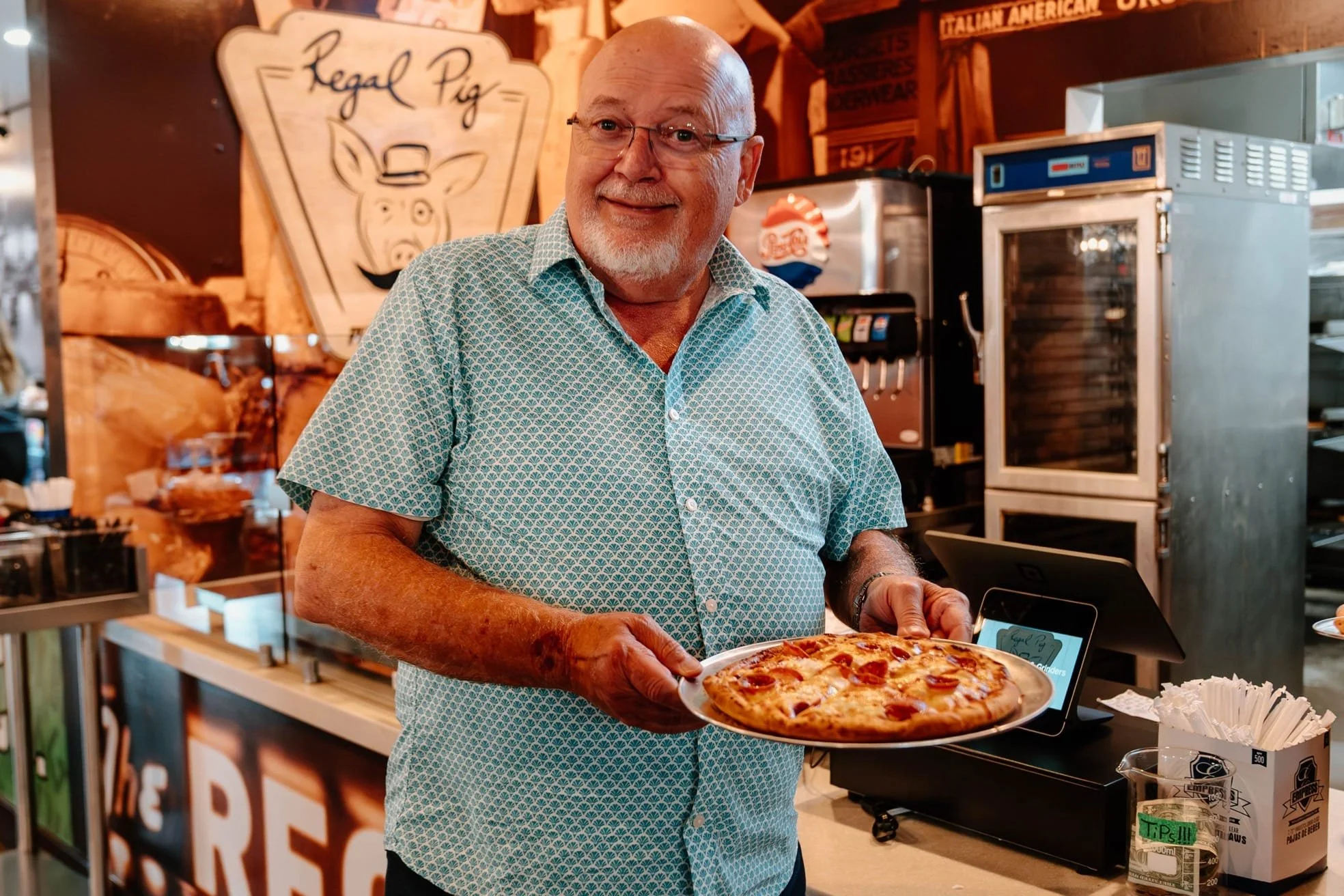 An older man with glasses, a beard, and a patterned short-sleeve shirt, smiling and holding a pizza at a pizzeria.
