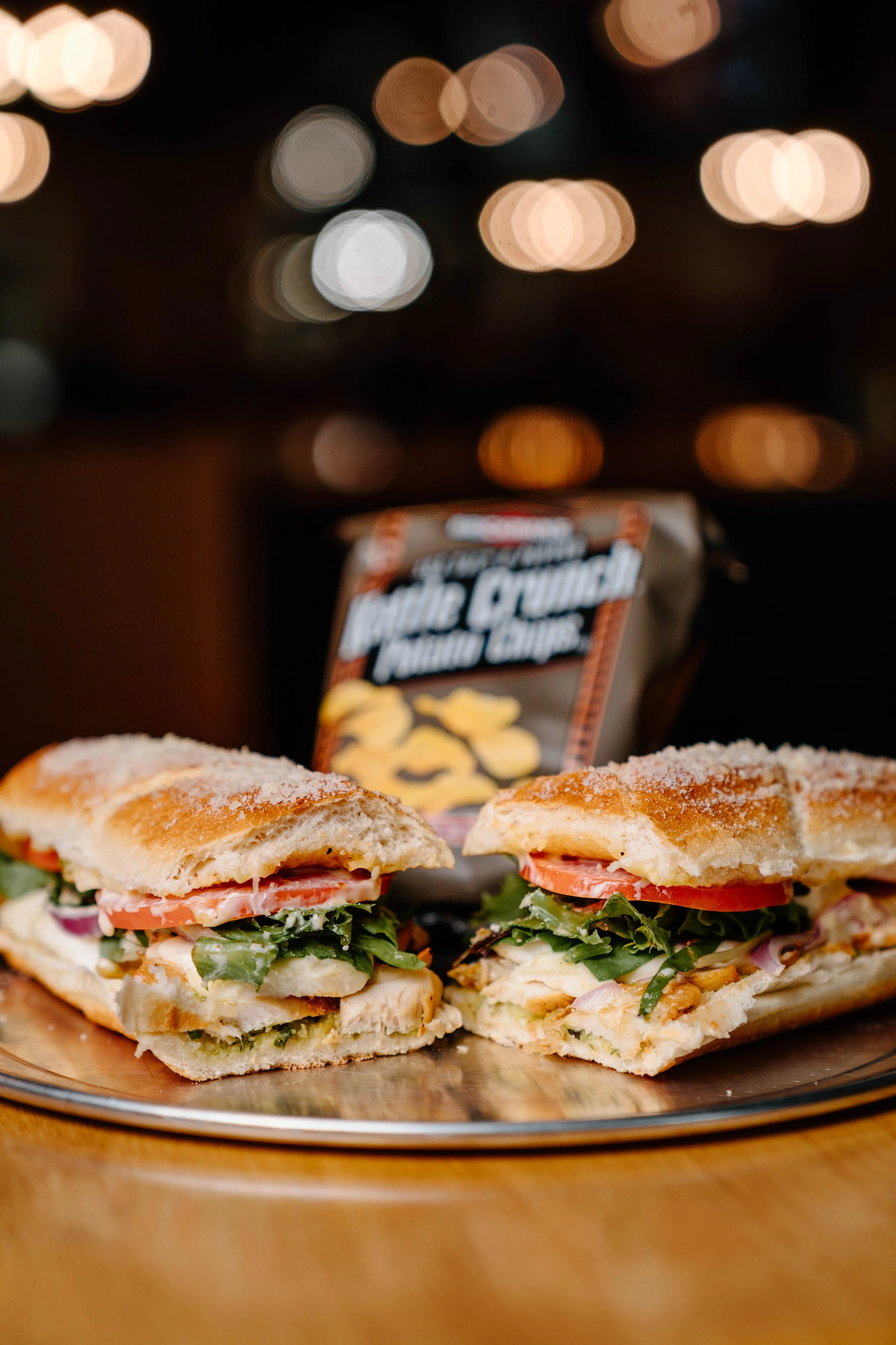 Close-up of a club sandwich with lettuce, tomato, meat, cheese, and condiments on toasted bread, on a glass plate, with a bag of potato chips in the background, and blurred lights behind.