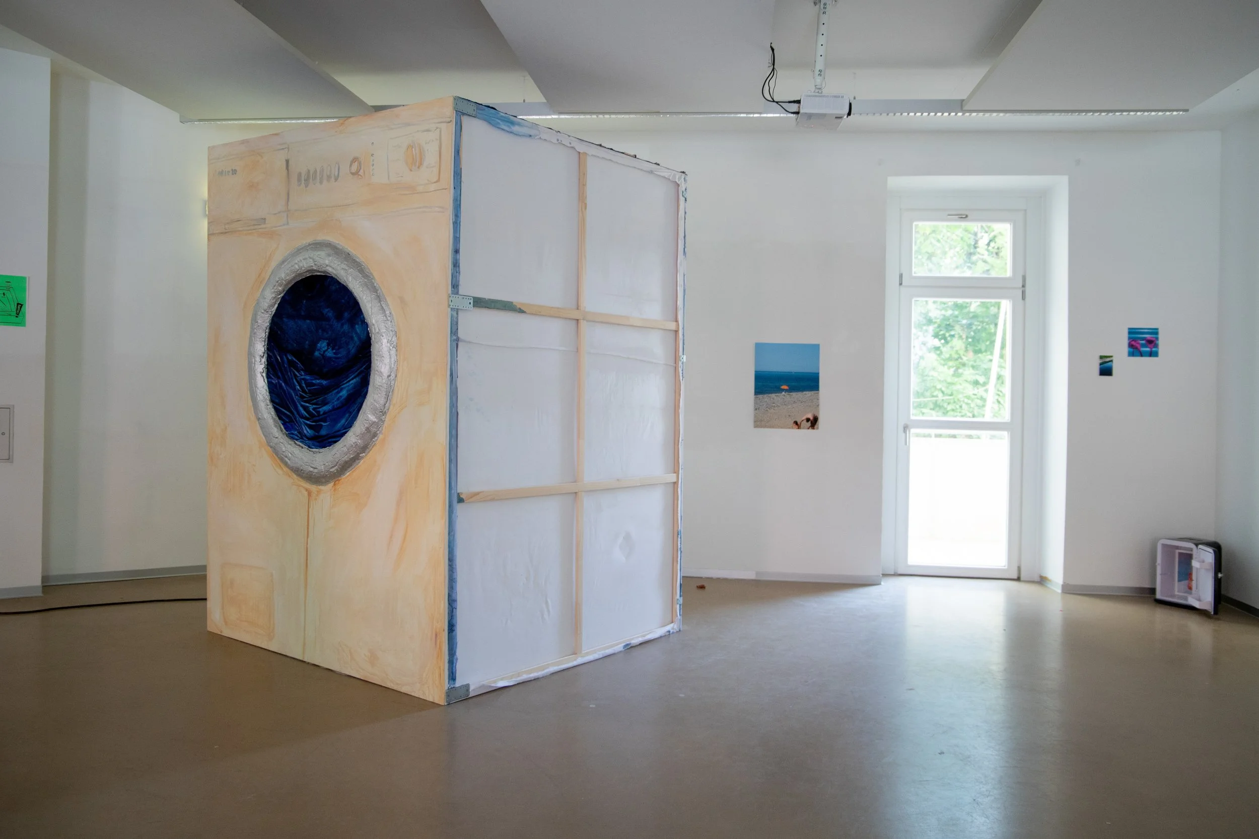 Kunstinstallation, die eine Waschmaschine nachahmt, in einem weißen Raum mit Fenstern, Wandbildern und einem schwarzen Gerät auf dem Boden.