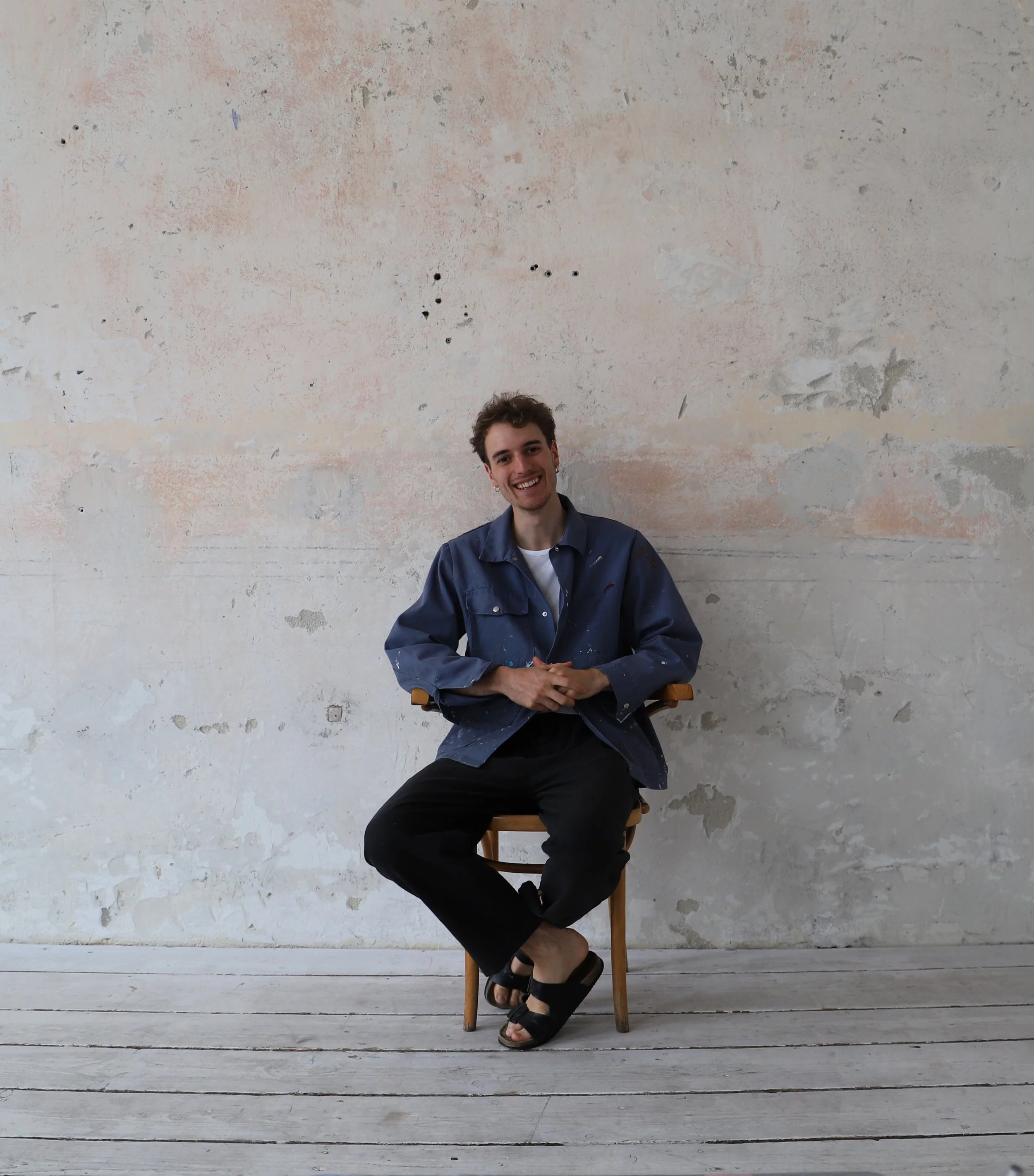 Clemens Gächter sitzt lächelnd auf einem Holzstuhl vor einer alten Wand mit pastellfarbenem Putz, trägt eine blaue Jacke, schwarze Hosen und Sandalen.