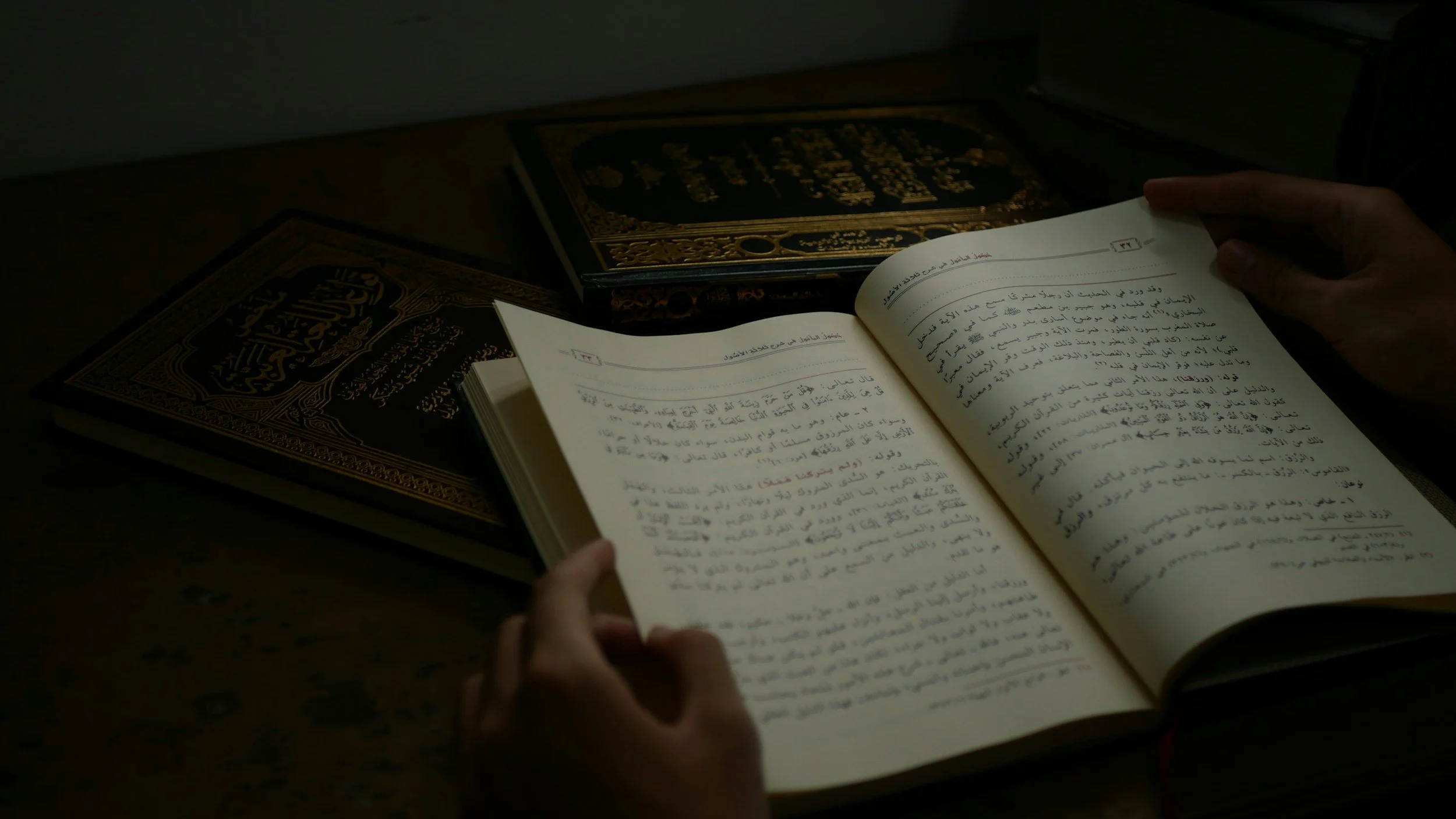 A person is reading an open book written in Arabic, with two other books with ornate black and gold covers placed on a dark wooden surface. The scene is dimly lit, focusing on the open book and hands.