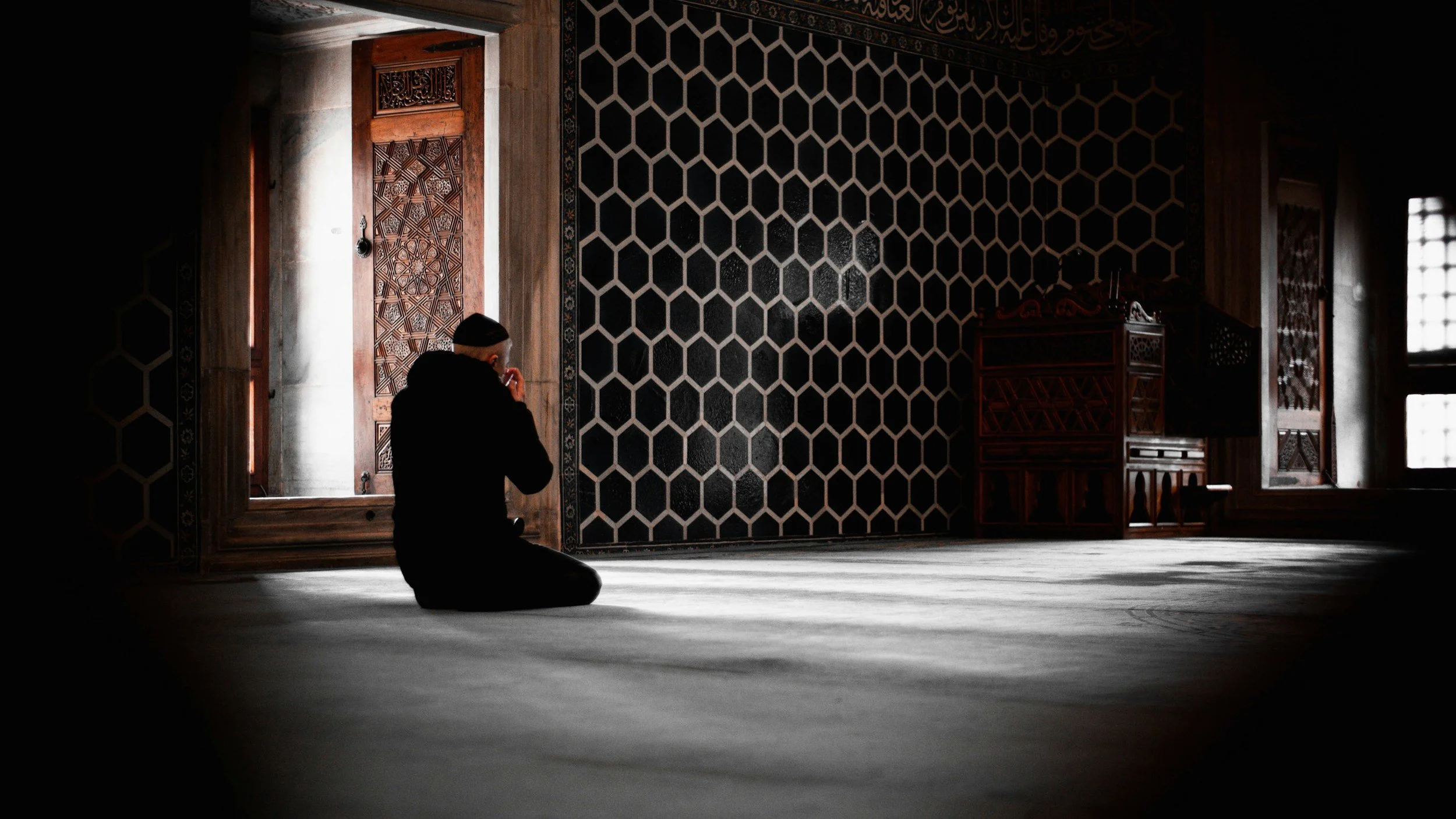 A person kneeling and praying inside a mosque, sitting on the floor with their hands raised near their face. The mosque has intricate woodwork on the doors and furniture, and patterned black and white walls. Light streams through windows, creating a serene atmosphere.
