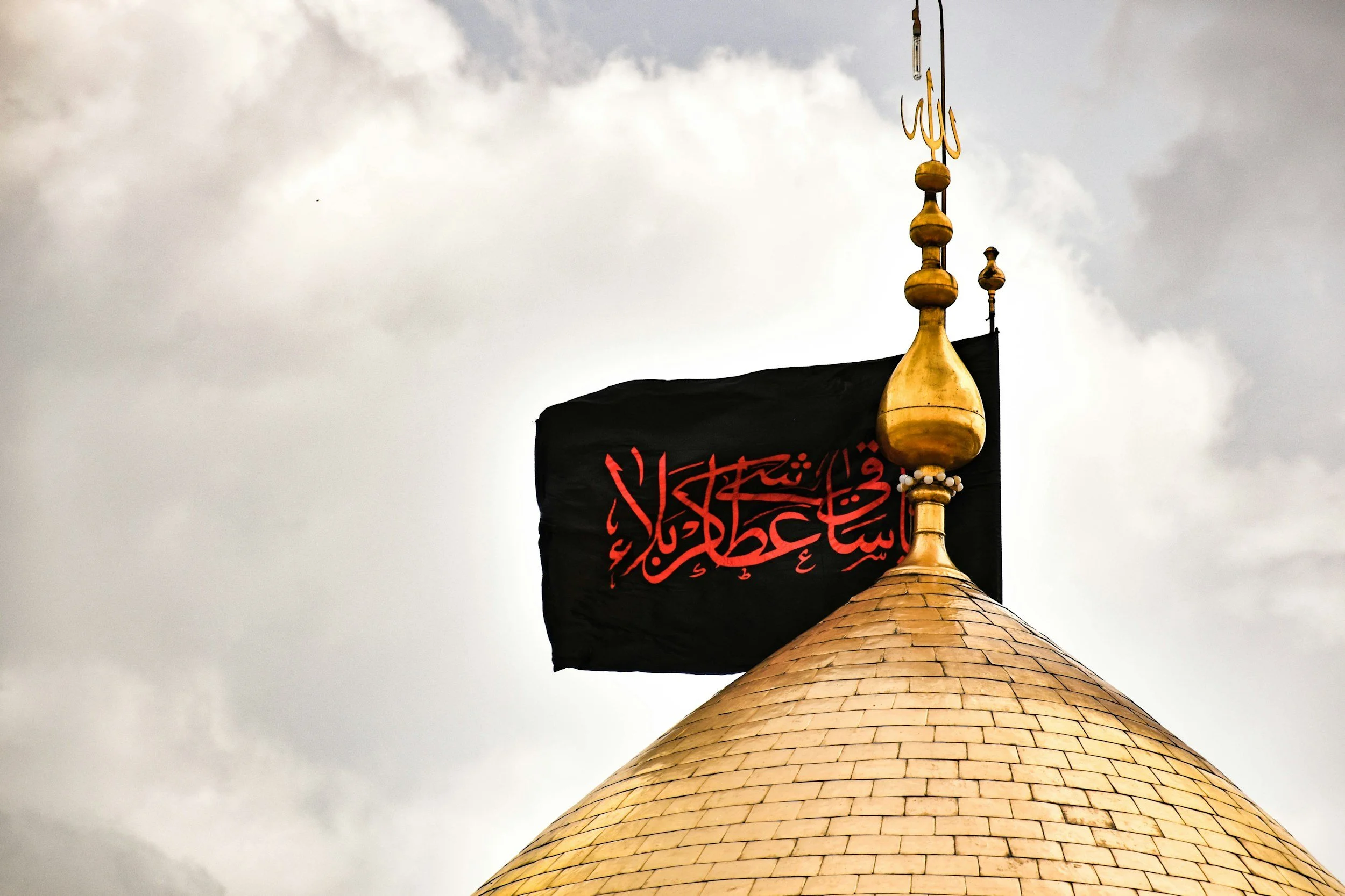 Islamic flag with Arabic script flying on top of a golden dome against cloudy sky.