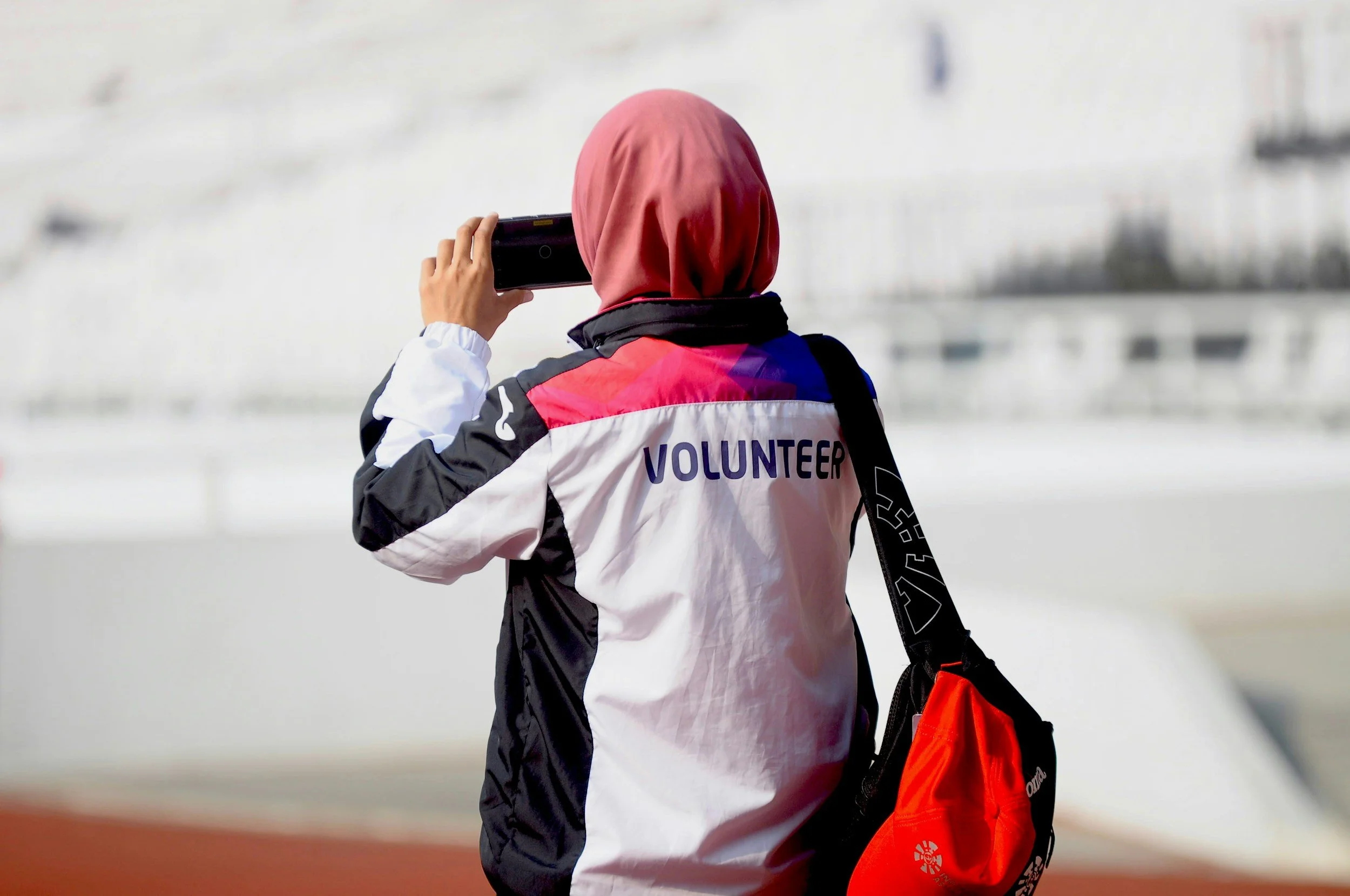 A person wearing a jacket labeled 'Volunteer' with a pink, purple, and white design, taking a photo with a smartphone. They have a red hijab and are carrying a black and red bag.