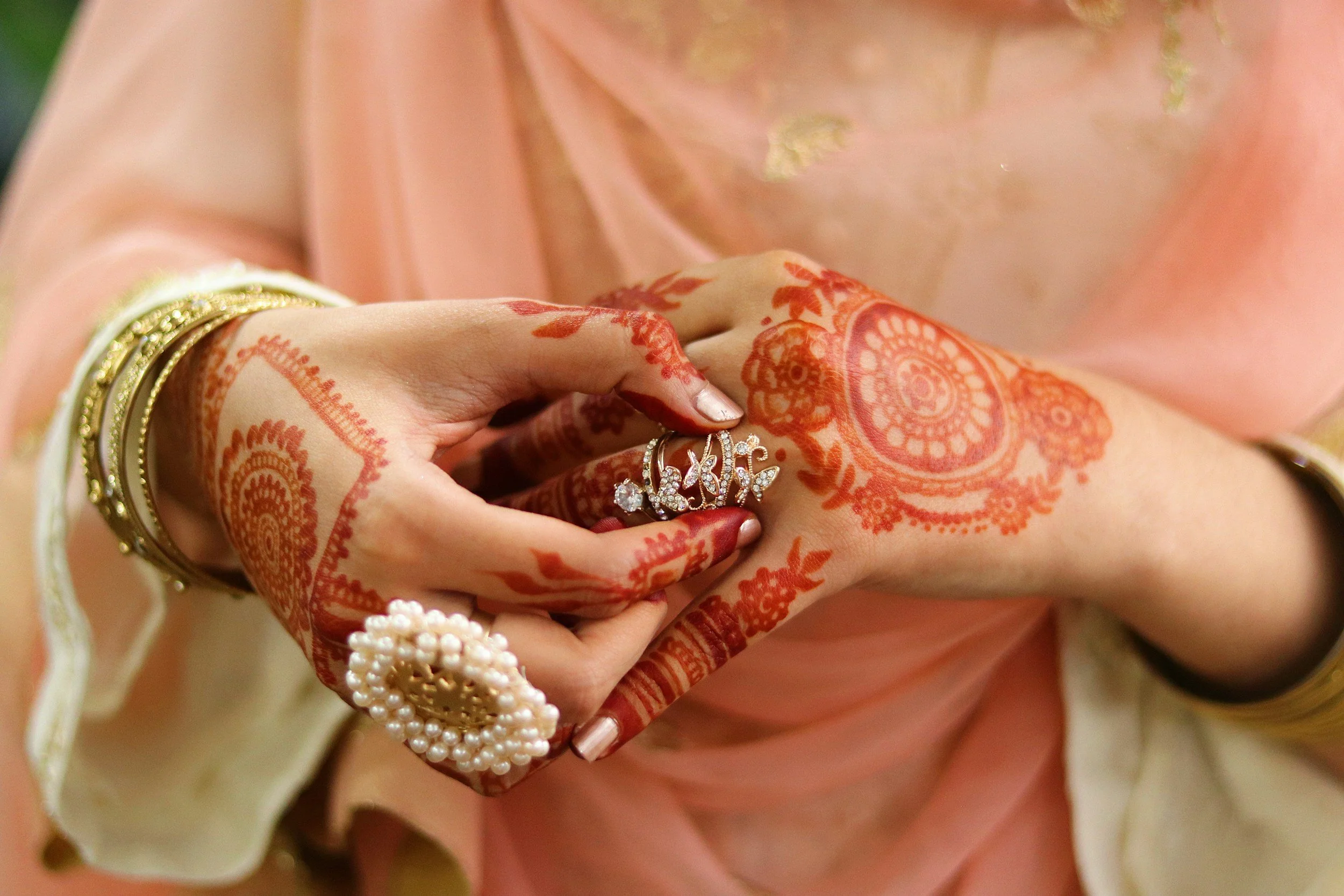 Close-up of hands with traditional henna designs, adorned with jewelry including bangles, a large ring with pearls, and a ring spelling 'Love,' wearing a peach-colored saree with gold embroidery.