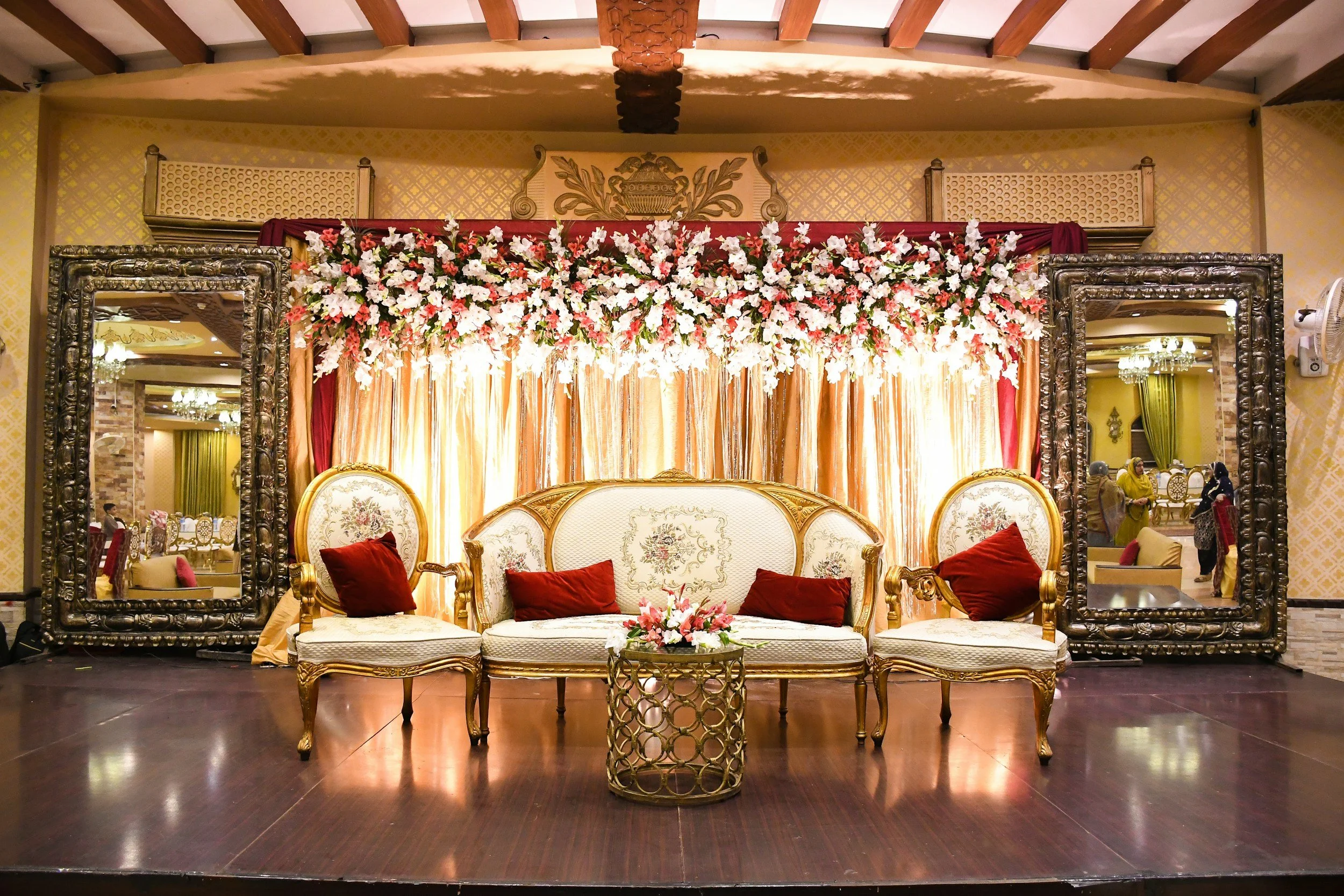 Elegant stage decorated with a floral backdrop featuring pink and white flowers, gold and white antique-style sofa with red pillows, mirrored frames on either side, and a small round table with a floral arrangement in front of the sofa.