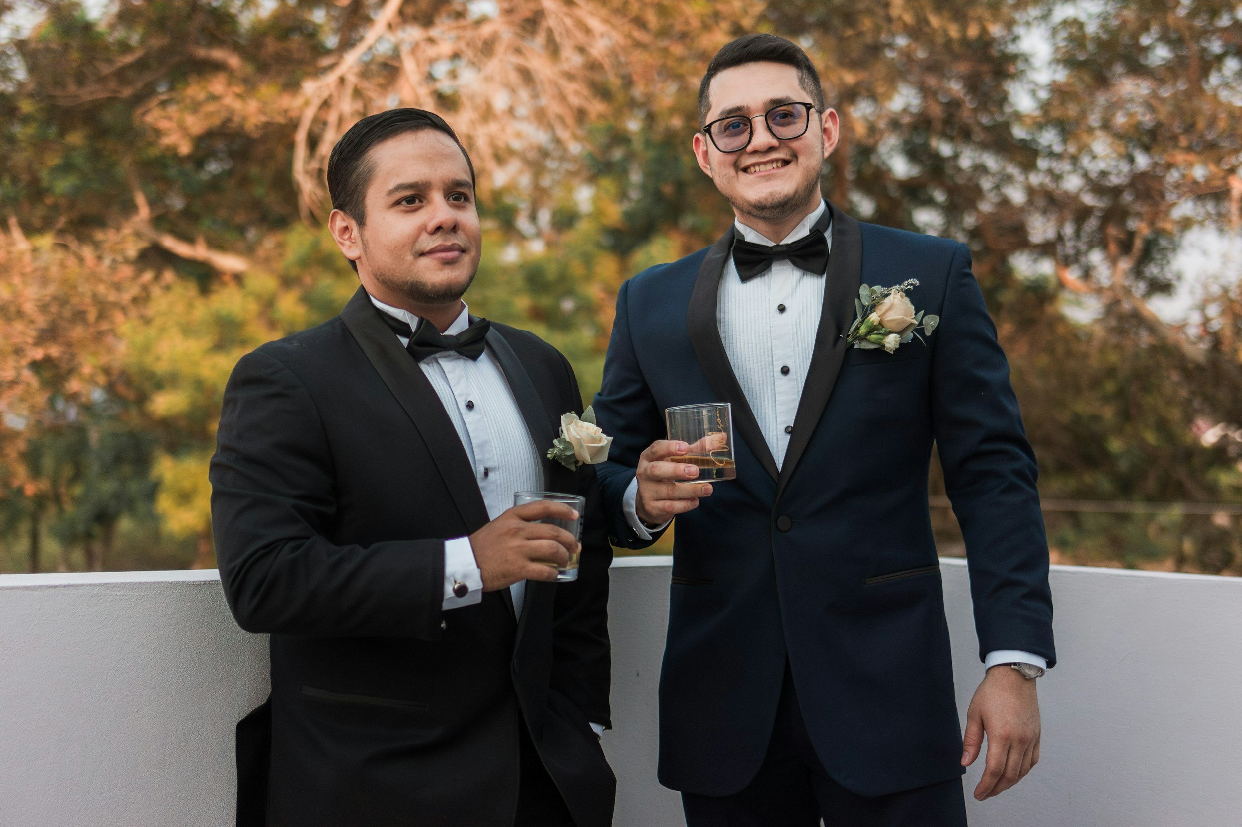 Two men in tuxedos with boutonnières, holding glasses of whiskey, standing outdoors on a balcony with trees and autumn foliage in the background, smiling and posing for a photo.