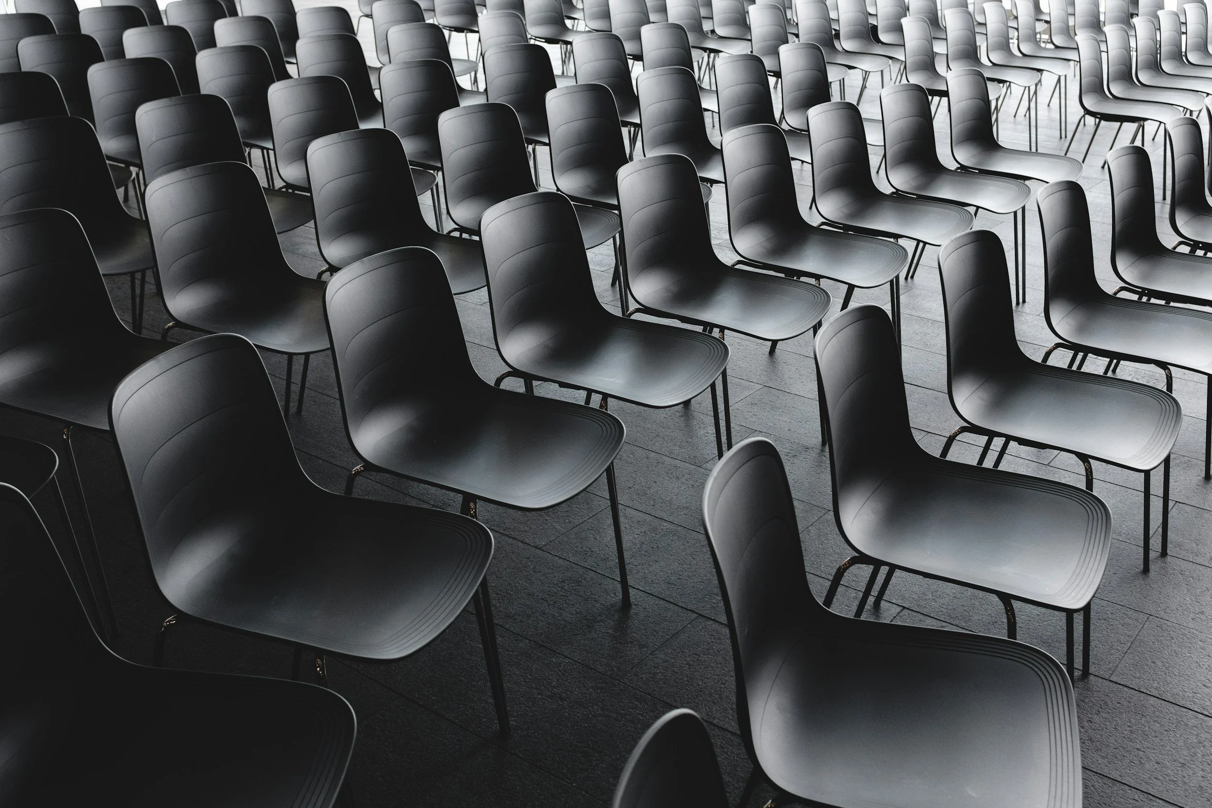 Multiple empty black chairs arranged in rows in a room with a grey wooden floor.