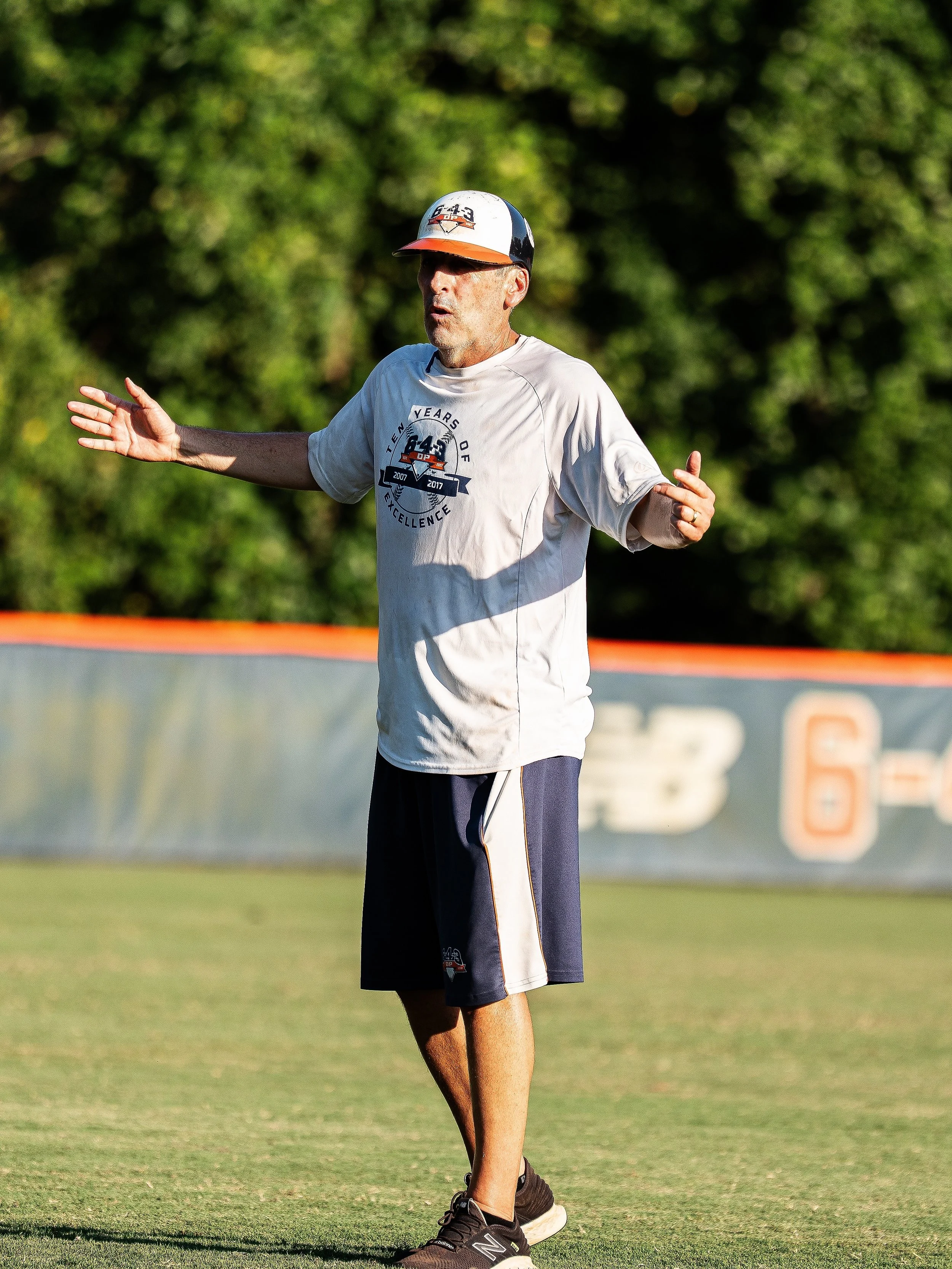A man in athletic clothing standing on a grass field, with a forest in the background, appearing to instruct or communicate during outdoor activity.