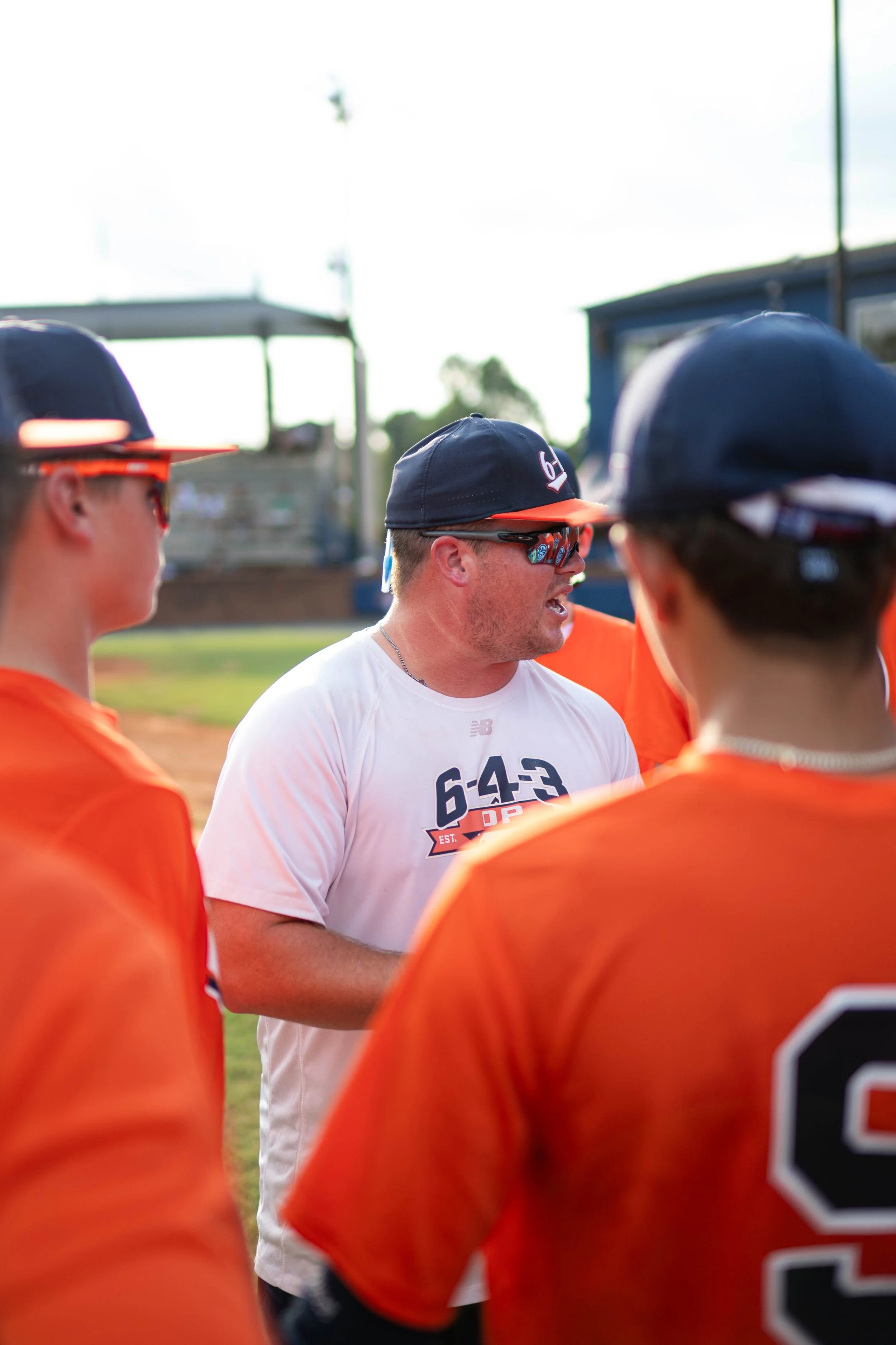 A coach speaking to a baseball team on the field, with players wearing orange uniforms and navy baseball caps, during late afternoon or early evening.