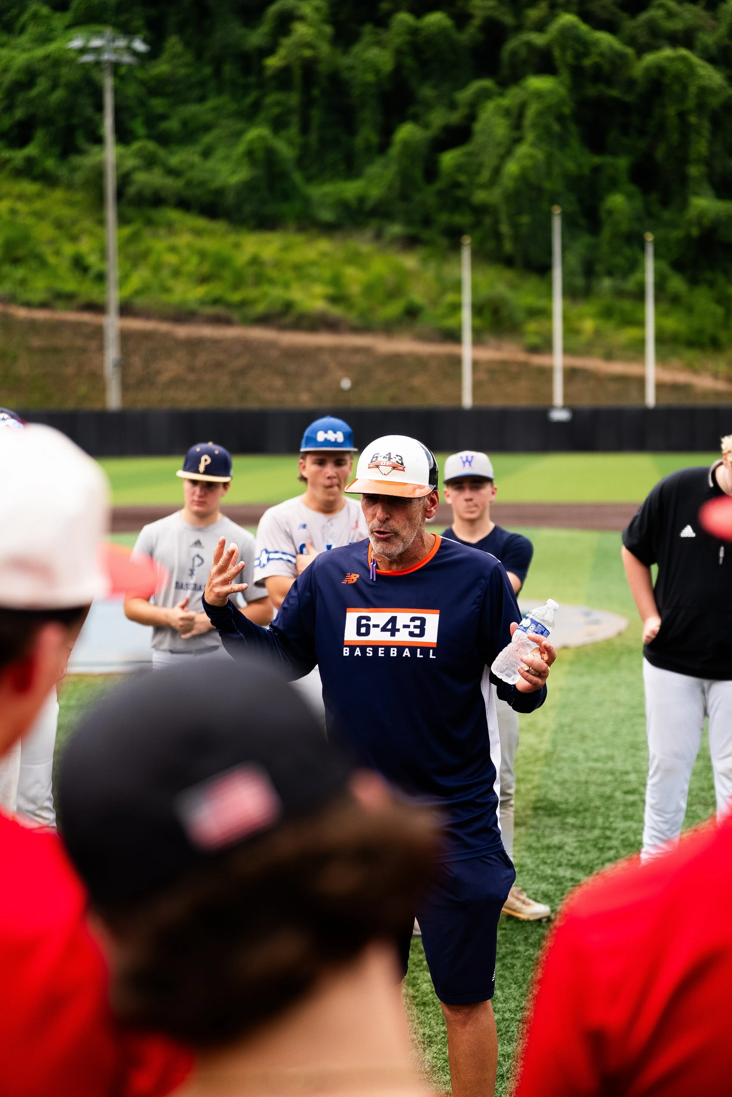 A baseball coach speaking to young athletes at a baseball field, with a backdrop of trees and a hill, during daytime.