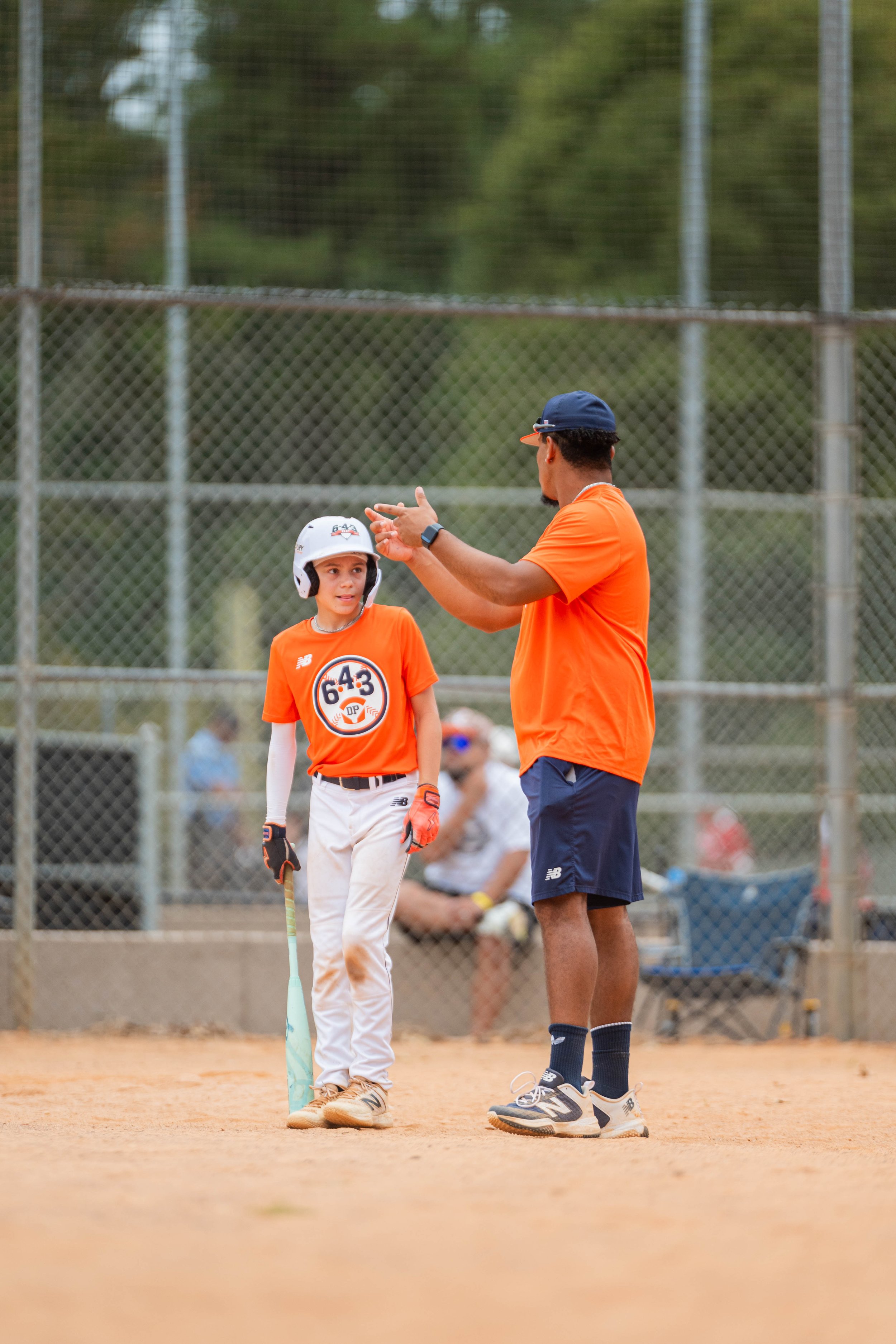 A coach teaching a young baseball player at a baseball field. The coach is explaining something, while the girl listens with her helmet and baseball bat in hand. The scene is set on a baseball diamond with a chain-link fence in the background and spectators watching.