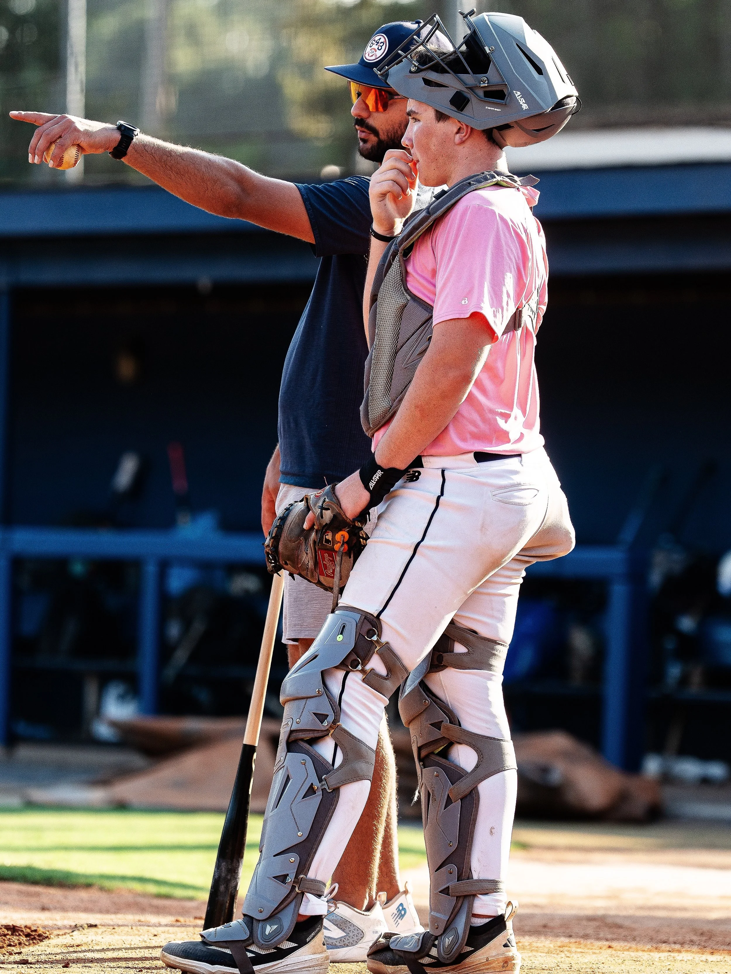 A baseball coach and a player standing on the baseball field, in conversation. The coach, wearing sunglasses and a cap, is pointing while the player, in catcher's gear including a helmet, chest protector, and shin guards, holds a baseball glove and appears to be listening attentively.