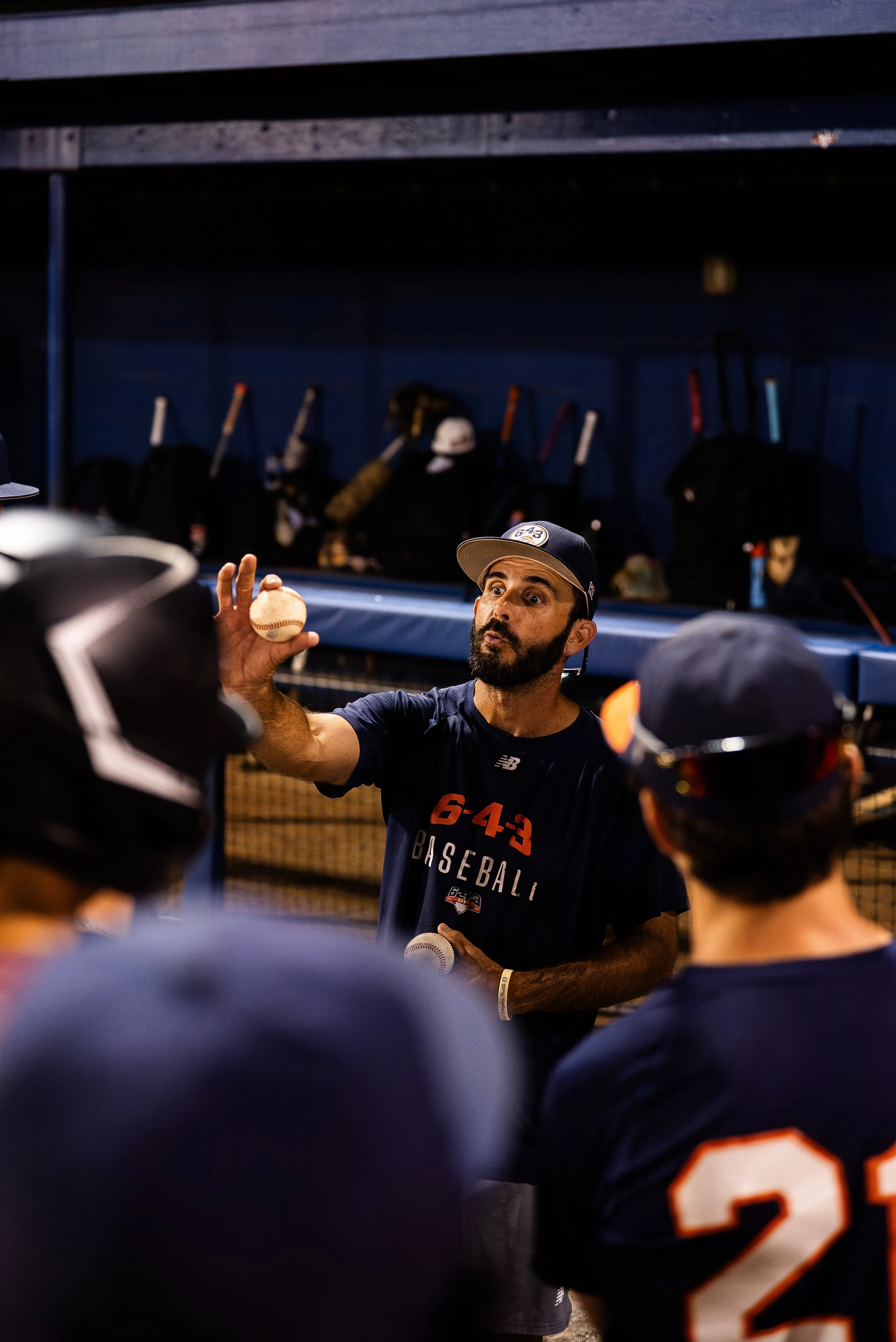 A baseball coach or trainer holding a baseball in a dugout or clubhouse, with players in baseball uniforms surrounding him. The coach appears to be giving instructions or motivation.