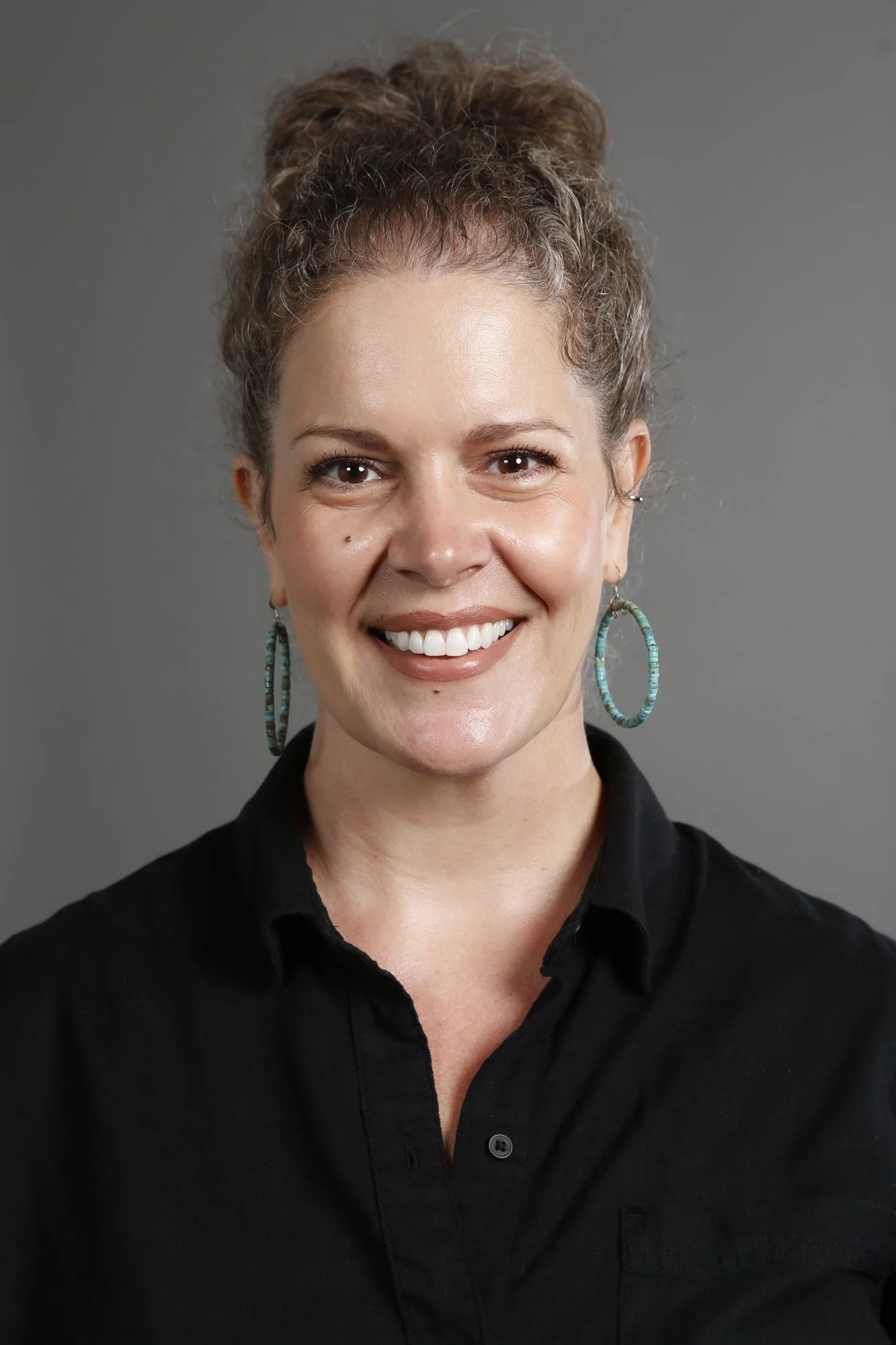 Kelli Monedero of Thinq is pictured with curly brown hair tied up, smiling, wearing turquoise hoop earrings and a black button-up shirt.
