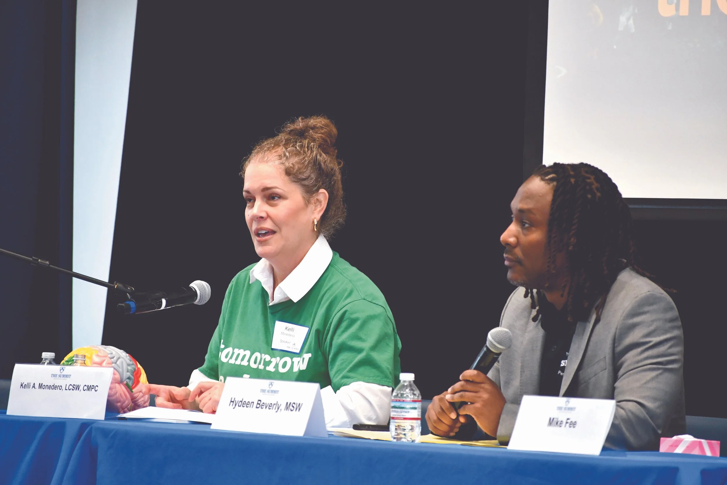 Two women sitting at a conference table with microphones. One woman is speaking, wearing a green shirt, with name tag Kelli A. Monedero. The other woman is listening, wearing a gray blazer, with name tag Mike Fee. A brain model and bottled water are on the table.