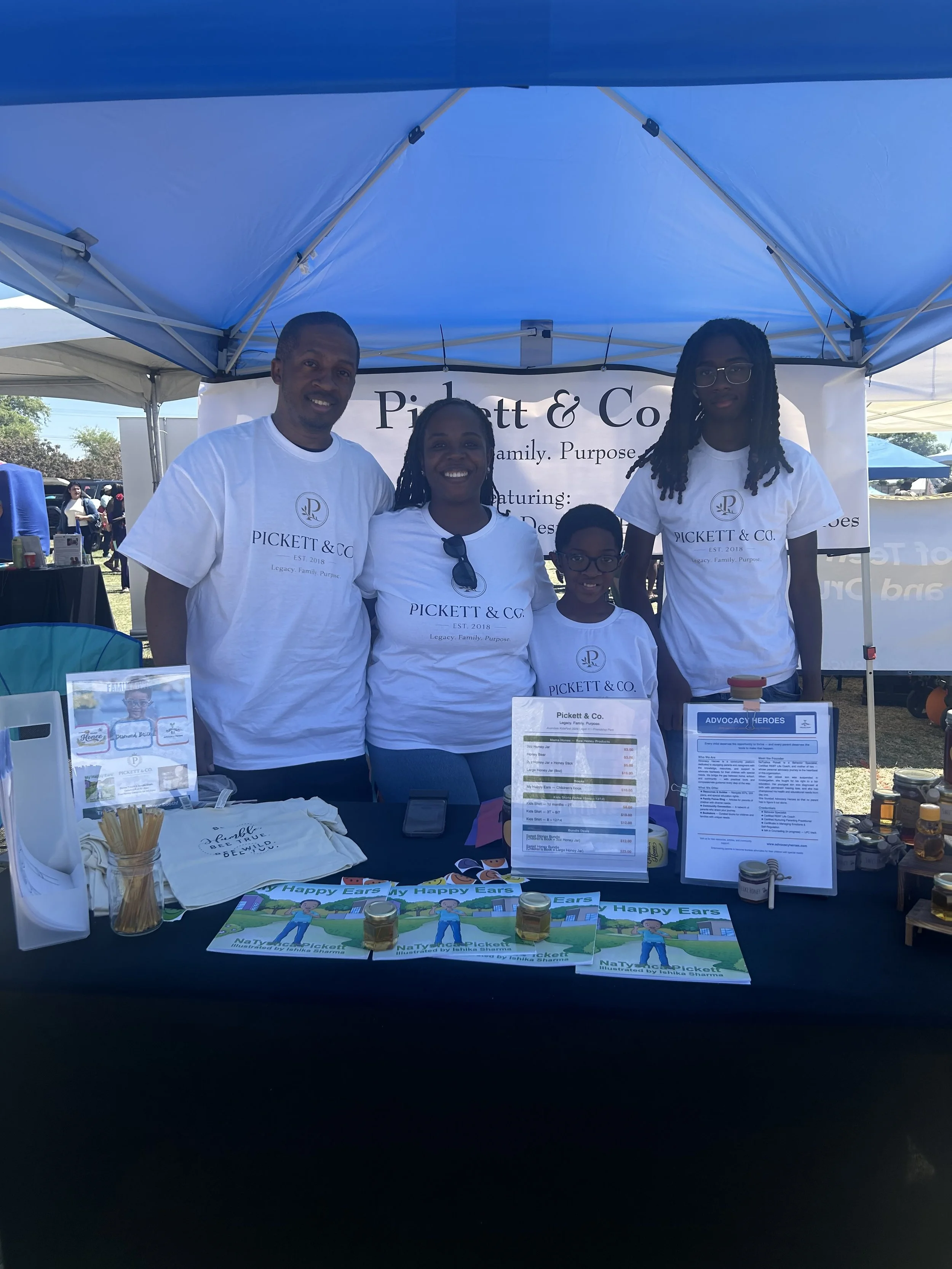 Family standing behind a booth at an outdoor event, displaying products and promotional materials for Pickett & Co.