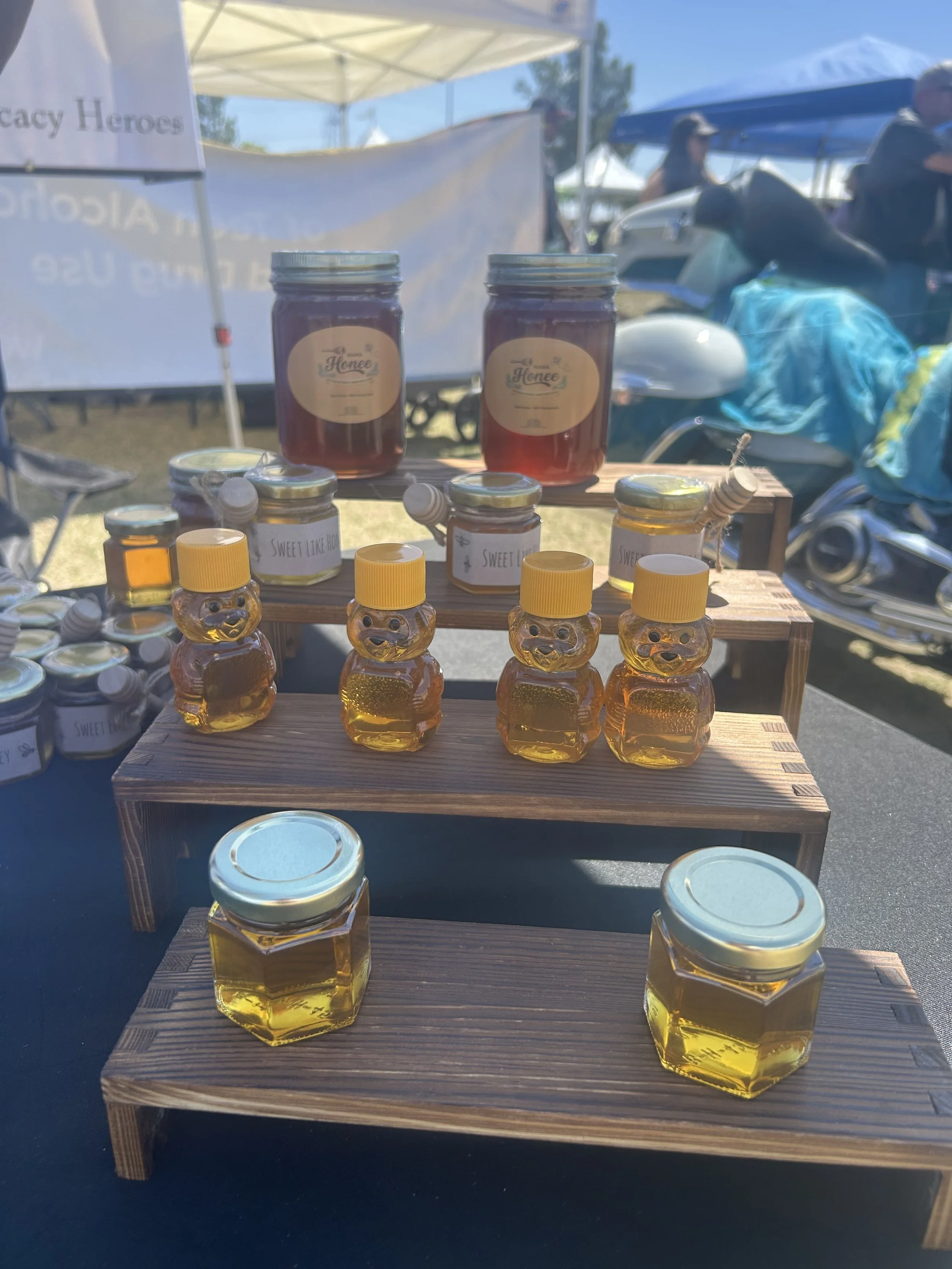 Display of honey jars and bear-shaped containers at an outdoor market stall, with tents and people in the background under a blue sky.