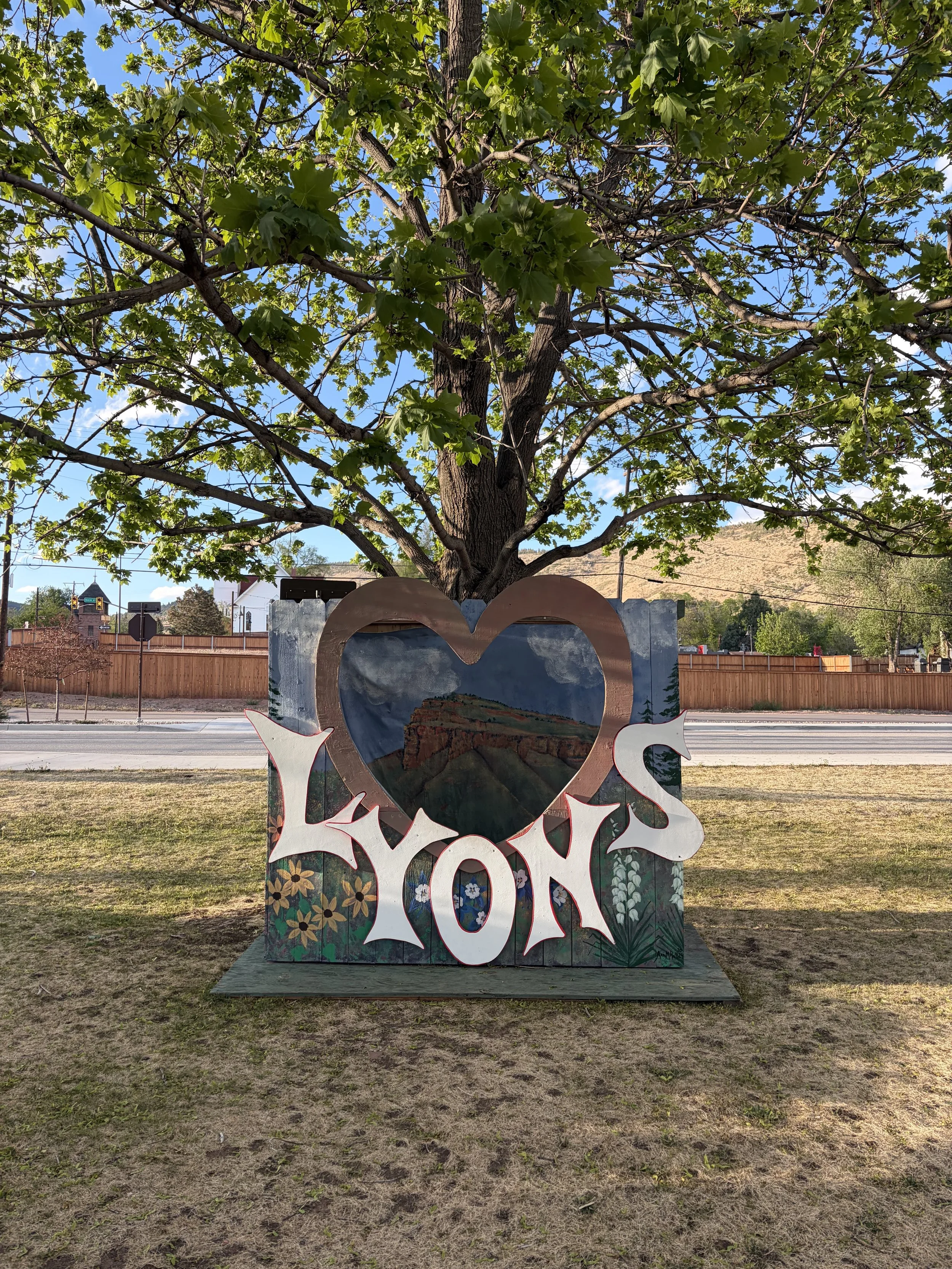 A decorative photo booth with the word 'LYONS' in white letters, a heart-shaped opening, and mountain patterns, placed outdoors in front of a large tree with green leaves and a blue sky.