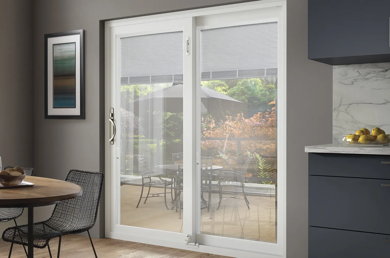 Sliding glass door leading to an outdoor patio with table and chairs, view of green trees and an umbrella outside, inside part of a kitchen and dining area with a framed picture on the wall and a bowl of lemons on the counter.