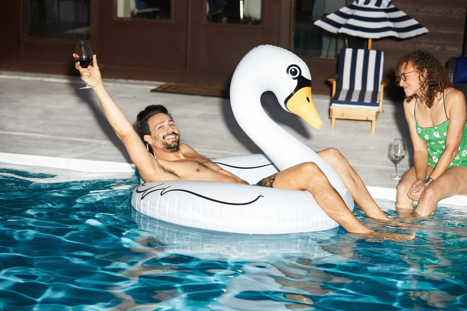 A man relaxes on a large white swan float in a swimming pool while a woman sits at the pool’s edge, both holding glasses of wine.