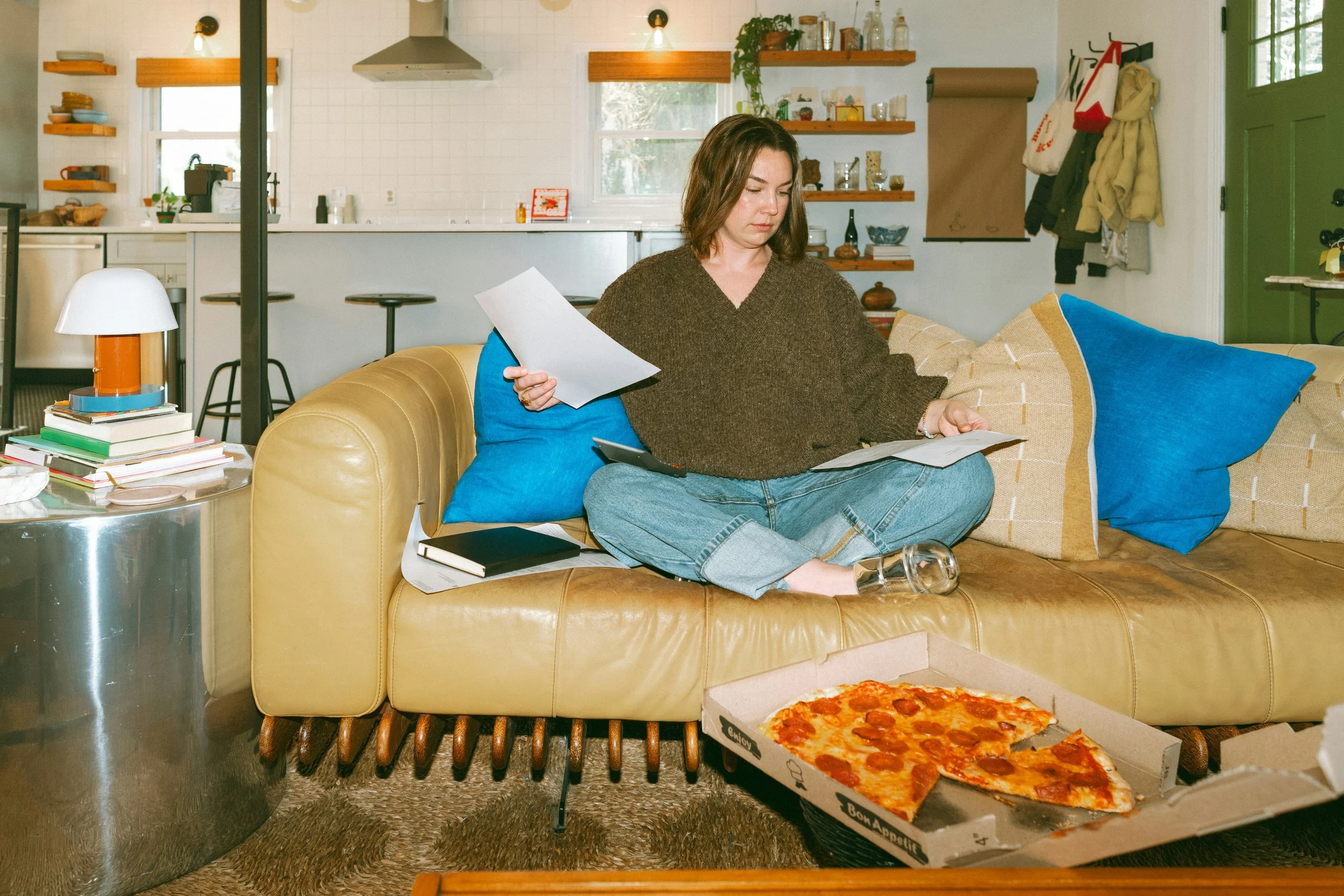 A woman sits cross-legged on a tan sofa sorting through papers, with bright blue cushions, an open kitchen behind her, and a half-eaten pizza on the coffee table.