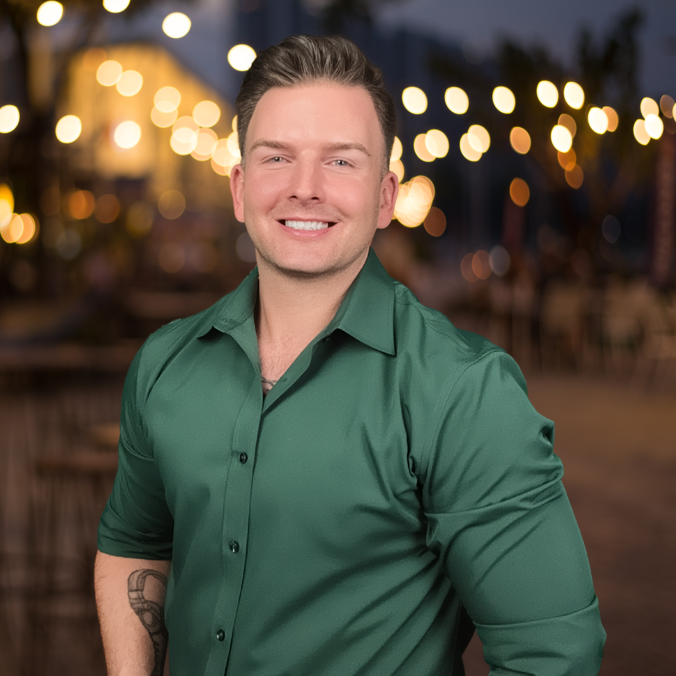 A young man with short dark hair, wearing a green button-up shirt, smiling, and standing outdoors at night with blurred string lights in the background.