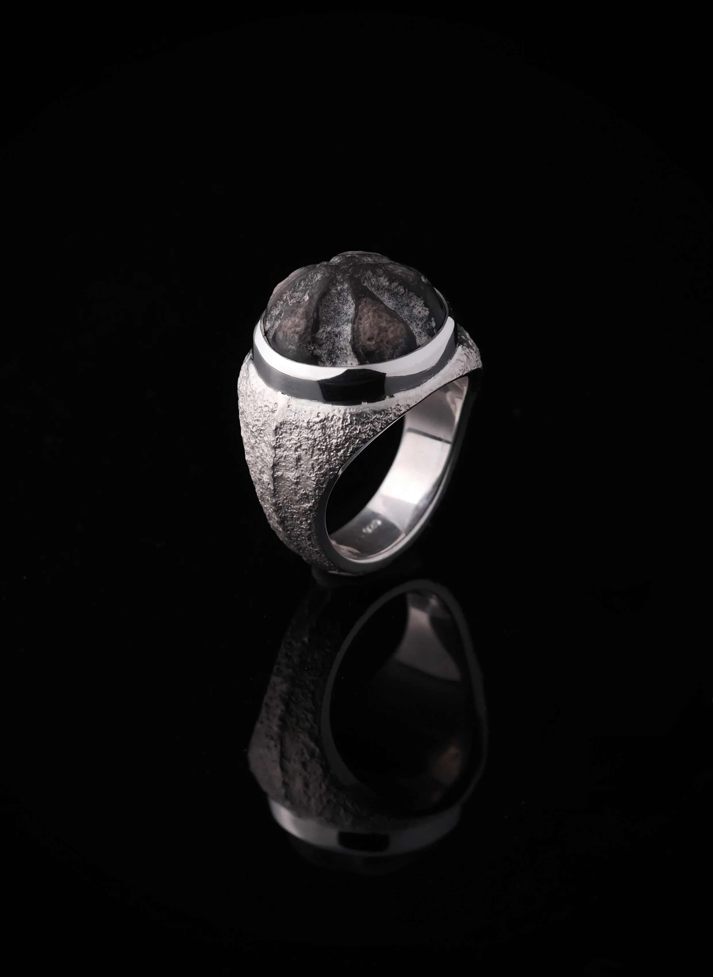 Ein silberner Ring mit großem, rauem, schwarzen Edelstein auf schwarzem Hintergrund, mit Reflexion auf der Oberfläche.