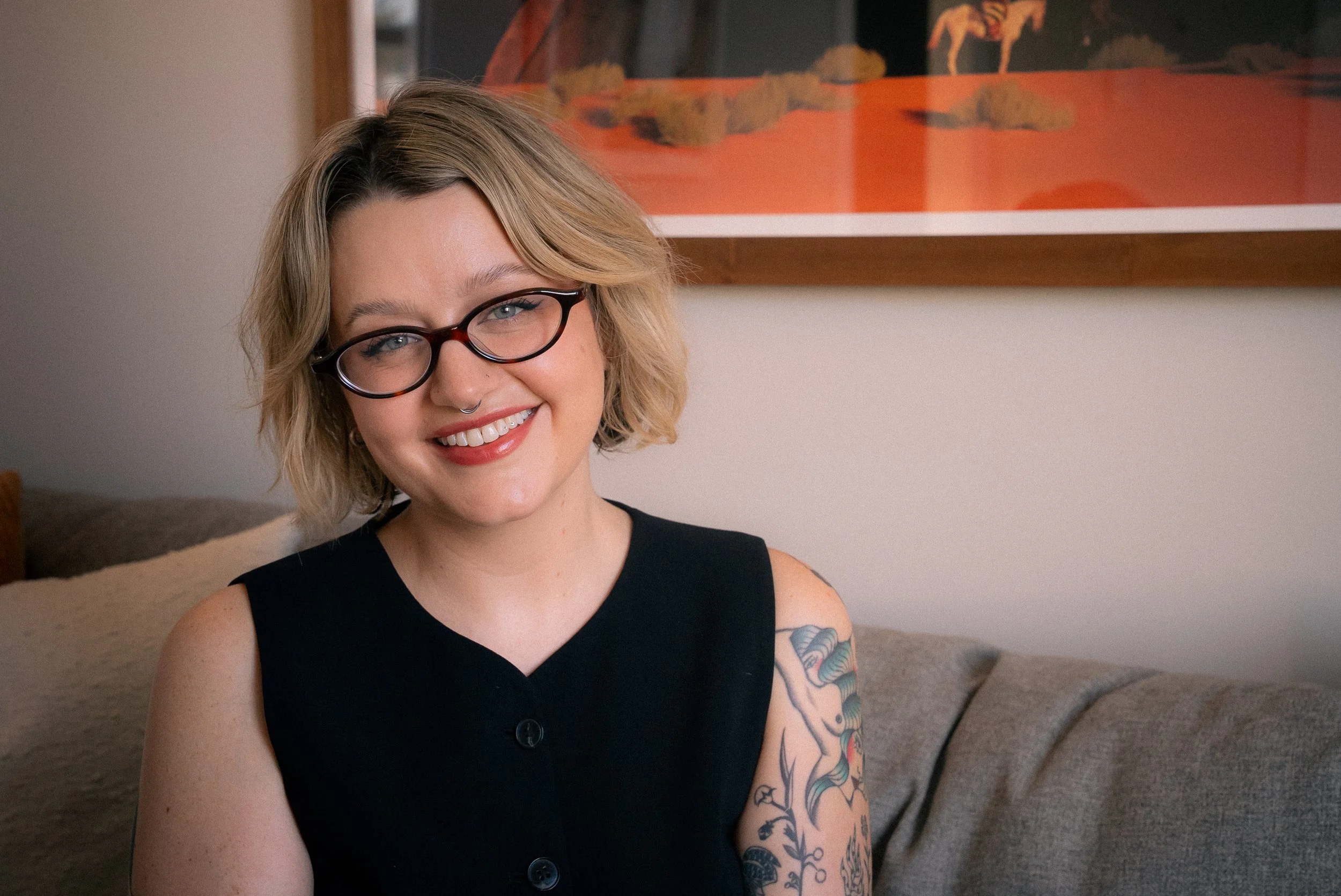 A woman with short blonde hair, glasses, and a septum piercing smiling while sitting on a couch, with a colorful framed artwork behind her.