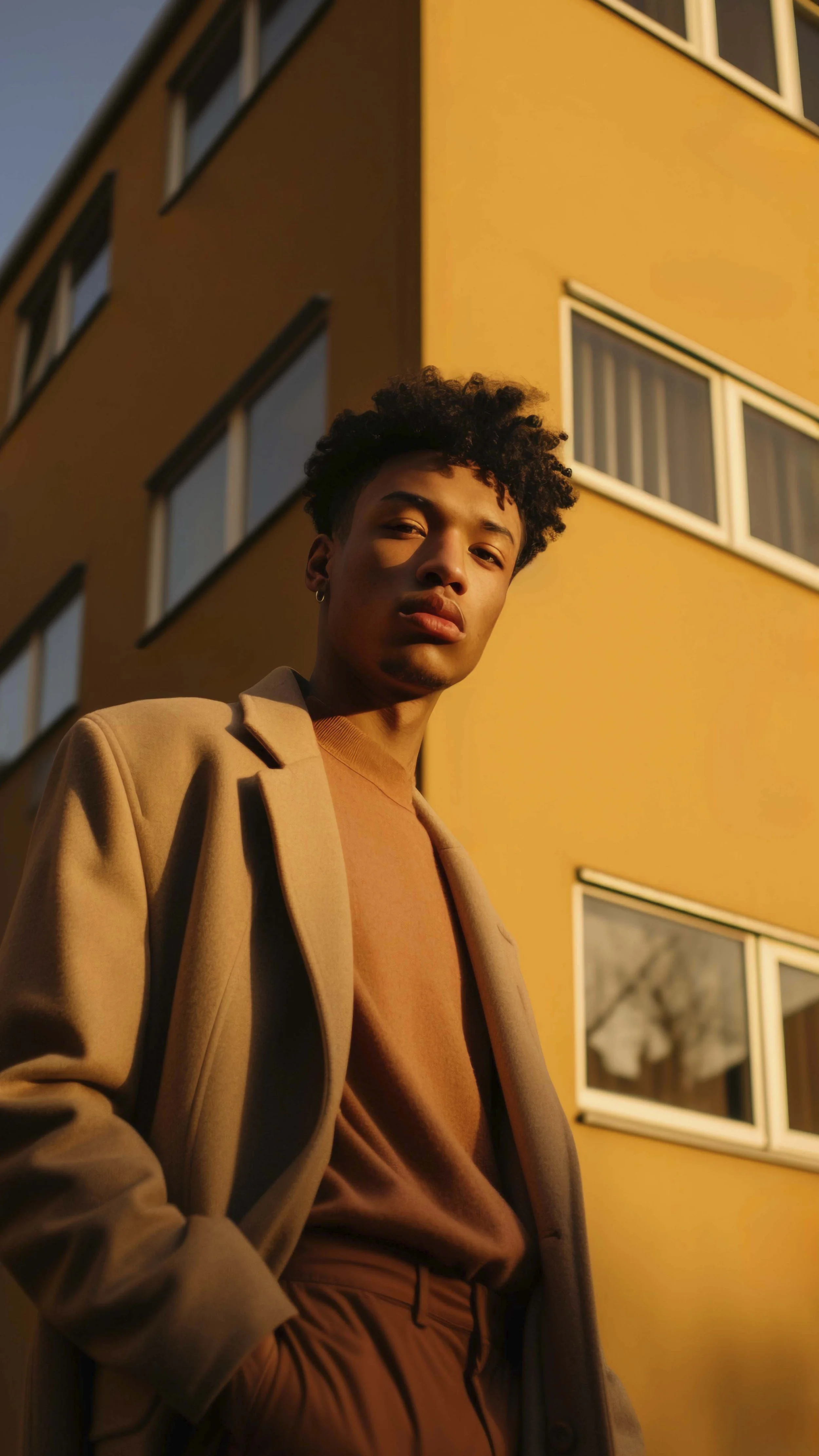 A young man with curly hair and earrings standing outdoors near a yellow building at sunset.