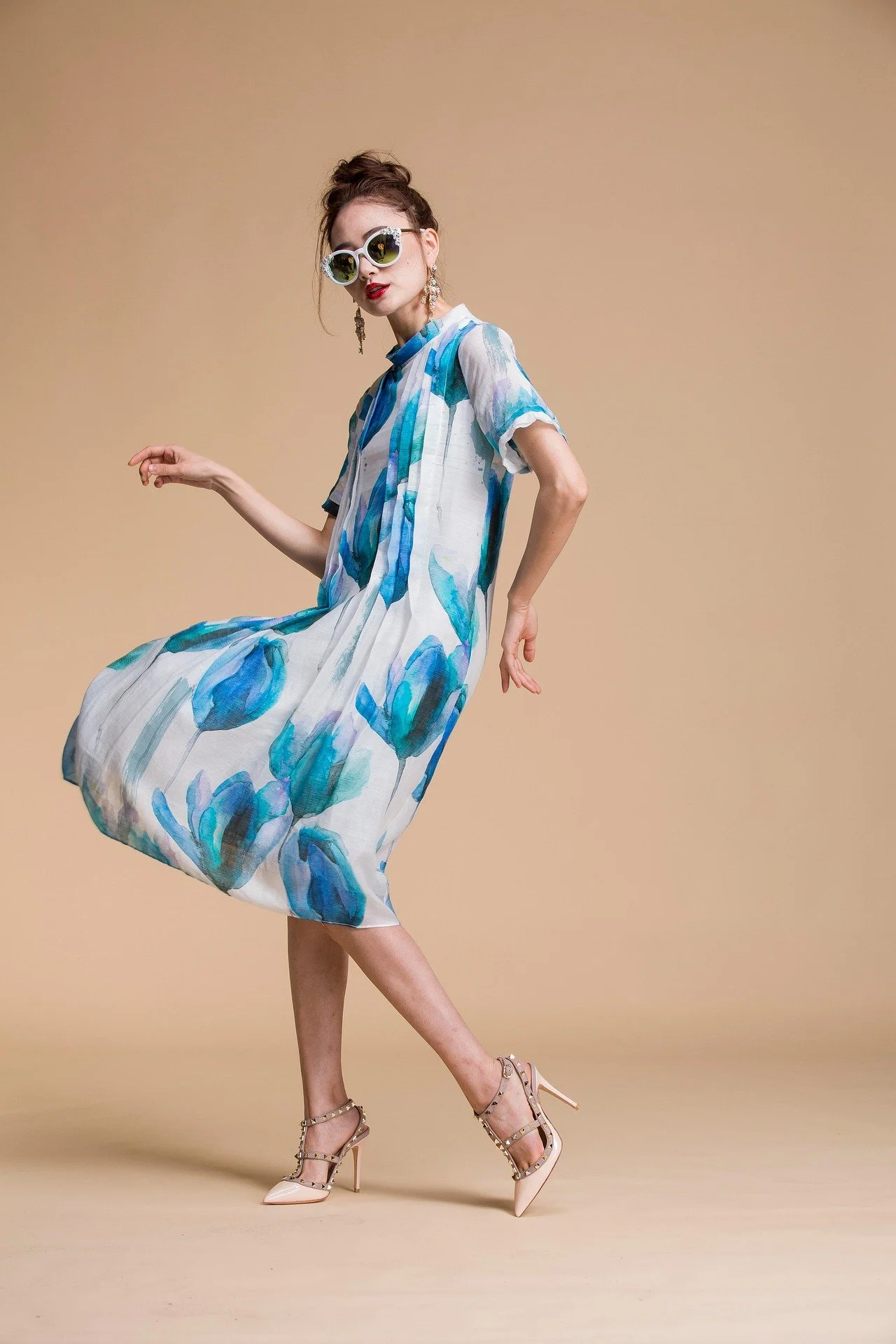 A woman wearing a flowy, knee-length dress with blue floral patterns, high-heeled shoes, large round sunglasses, and earrings, posing against a plain beige background.