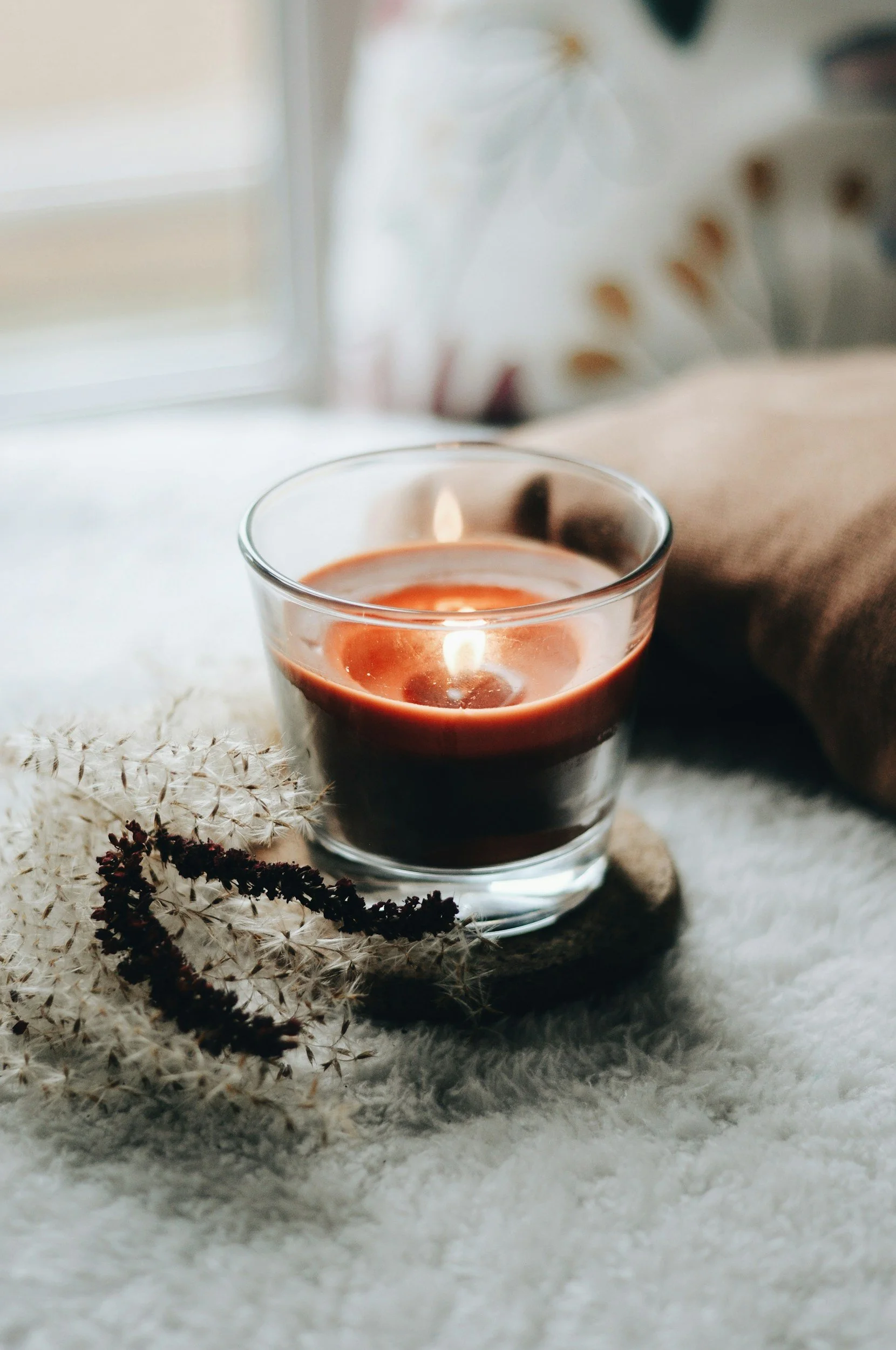 A glass containing a candle with a flickering flame, placed on a soft white surface along with dried plants, near a window with natural light