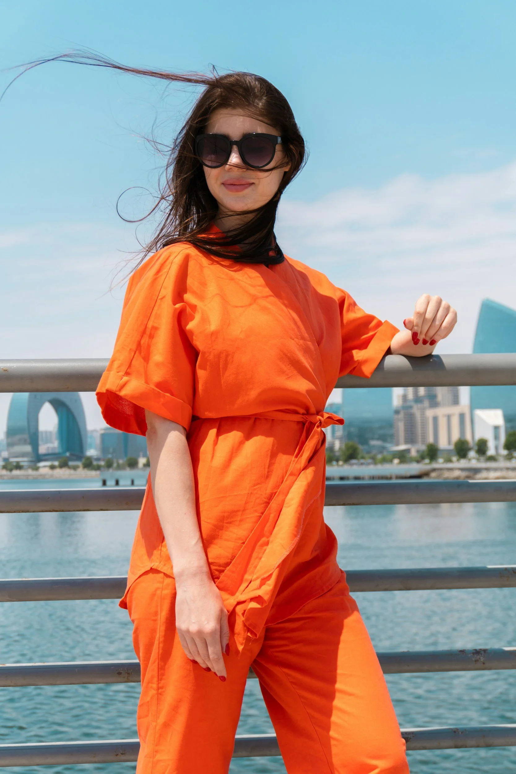 A woman wearing orange outfit and sunglasses standing by a metal railing with a cityscape and water behind her.