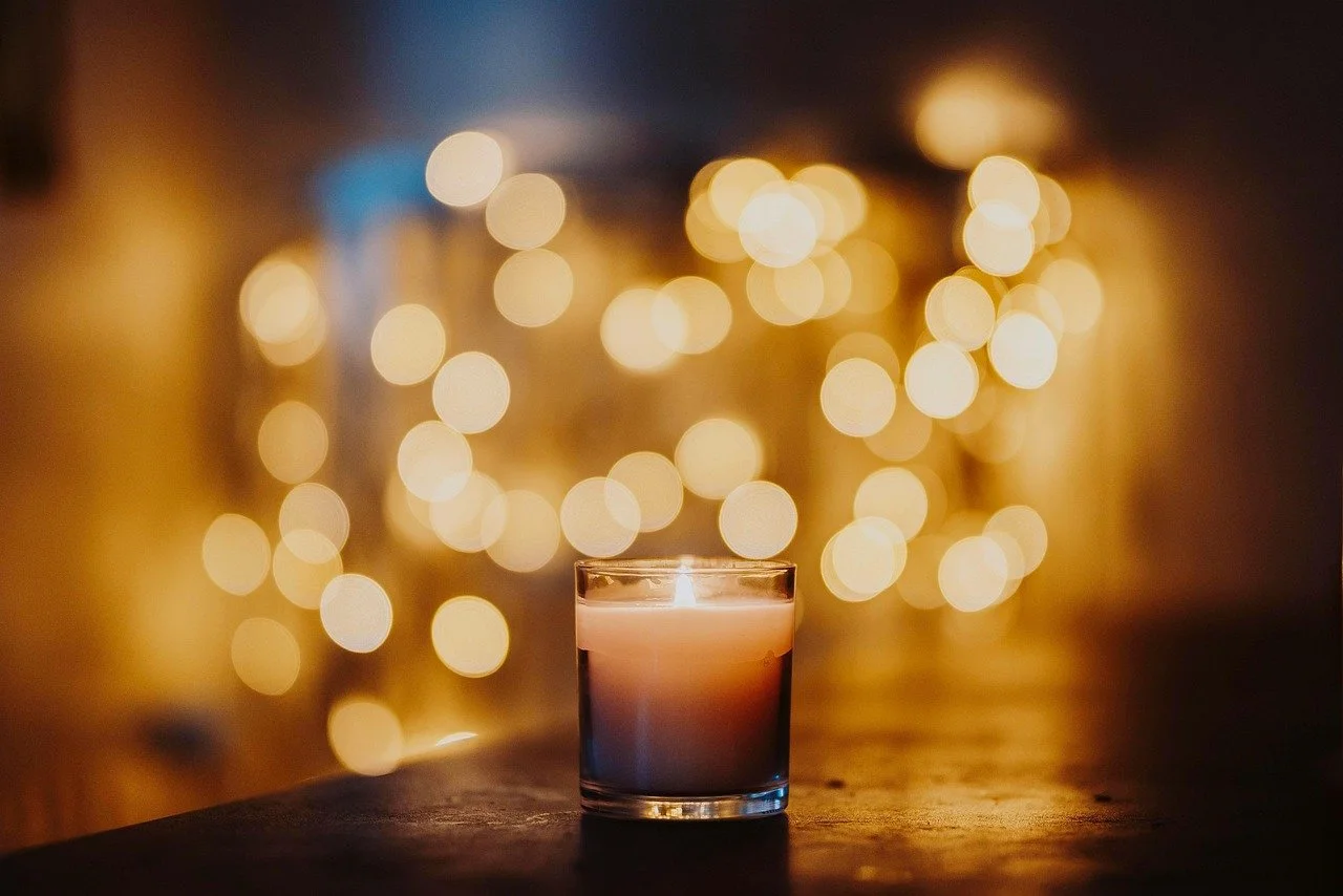 A lit candle in a glass holder on a wooden surface with blurred warm yellow lights in the background.
