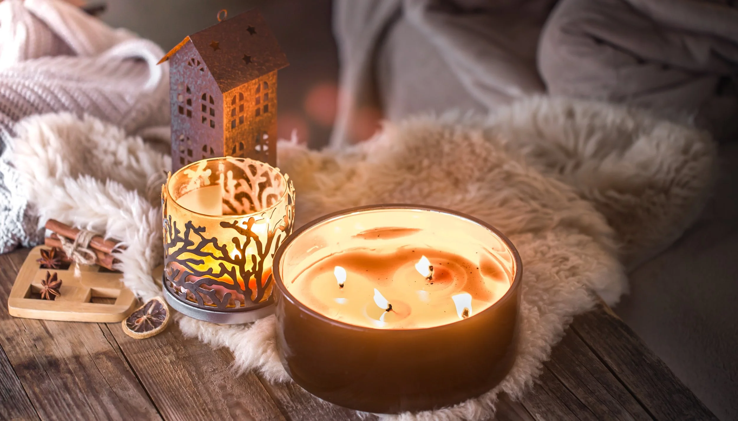 Cozy scene with lit candles, a small decorative house, star anise, cinnamon sticks, and a fleece blanket on a wooden surface.