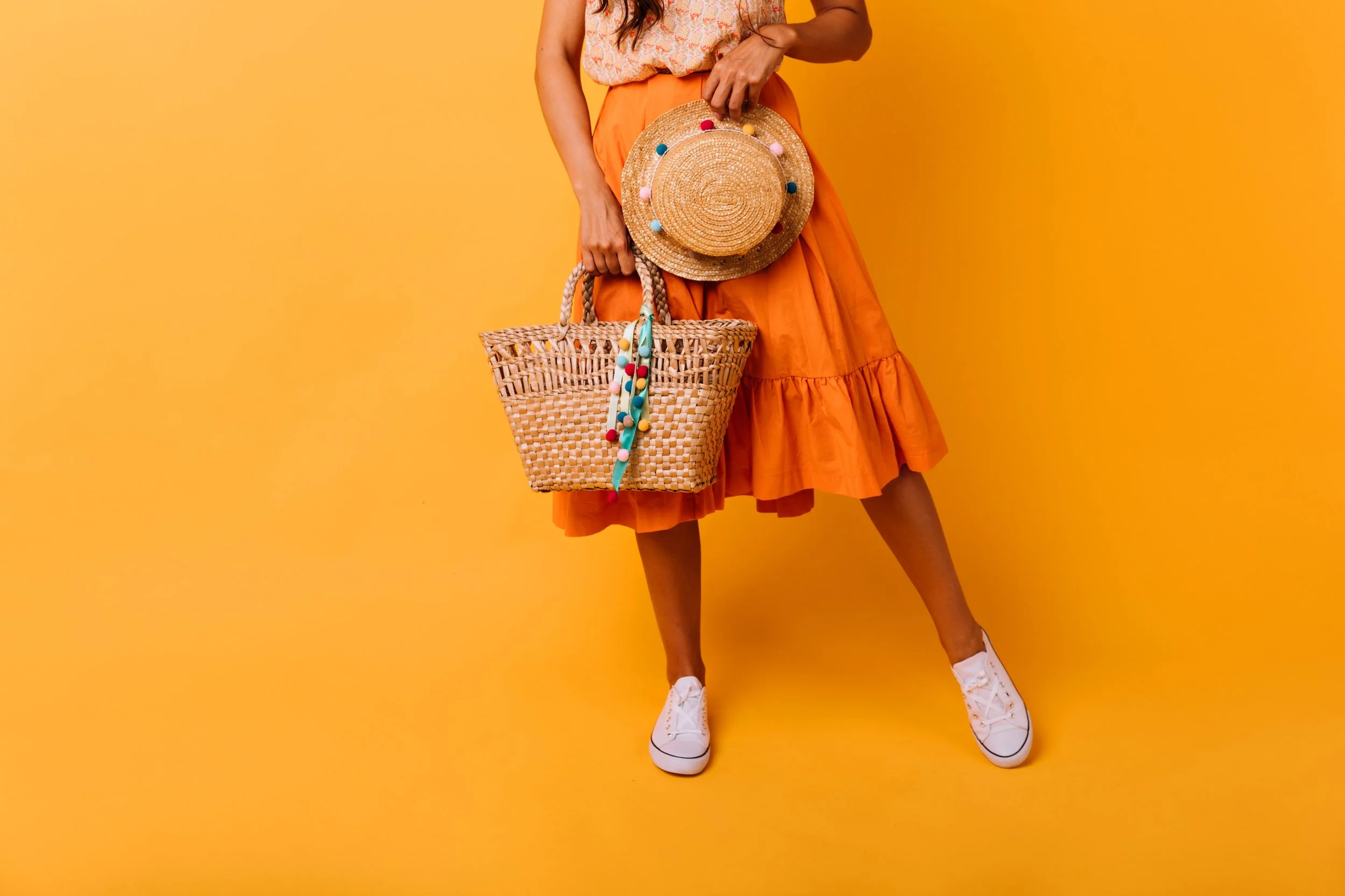 Person standing on yellow background wearing orange skirt, holding a woven straw handbag with colorful pom-pom accessories, and a wide-brimmed straw hat decorated with multicolored pompoms. White sneakers are visible.