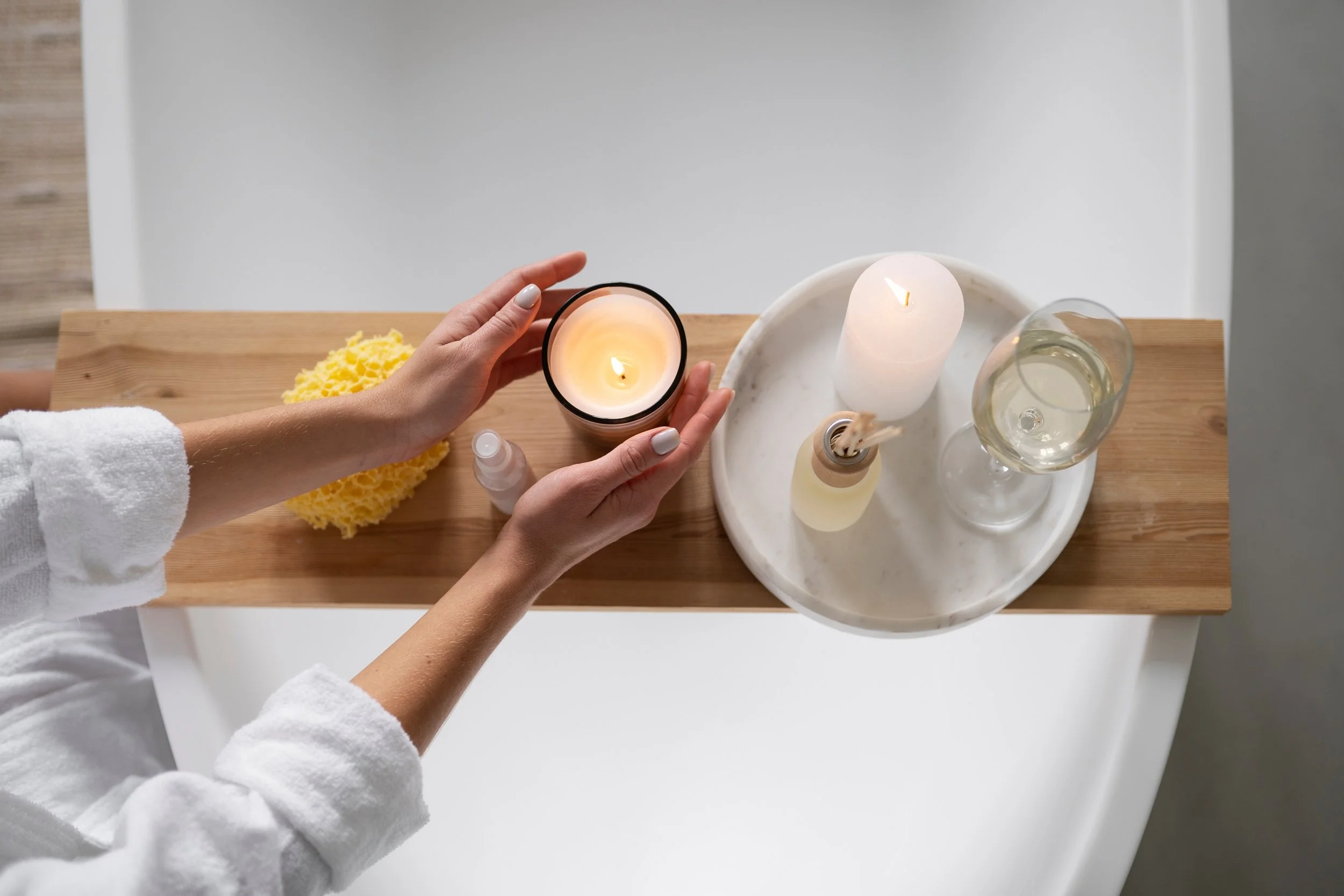 Top-down view of a person's hands holding a lit candle, with spa items on a wooden tray including another candle, a reed diffuser, a glass of white wine, and a yellow sponge, in a relaxing setting.
