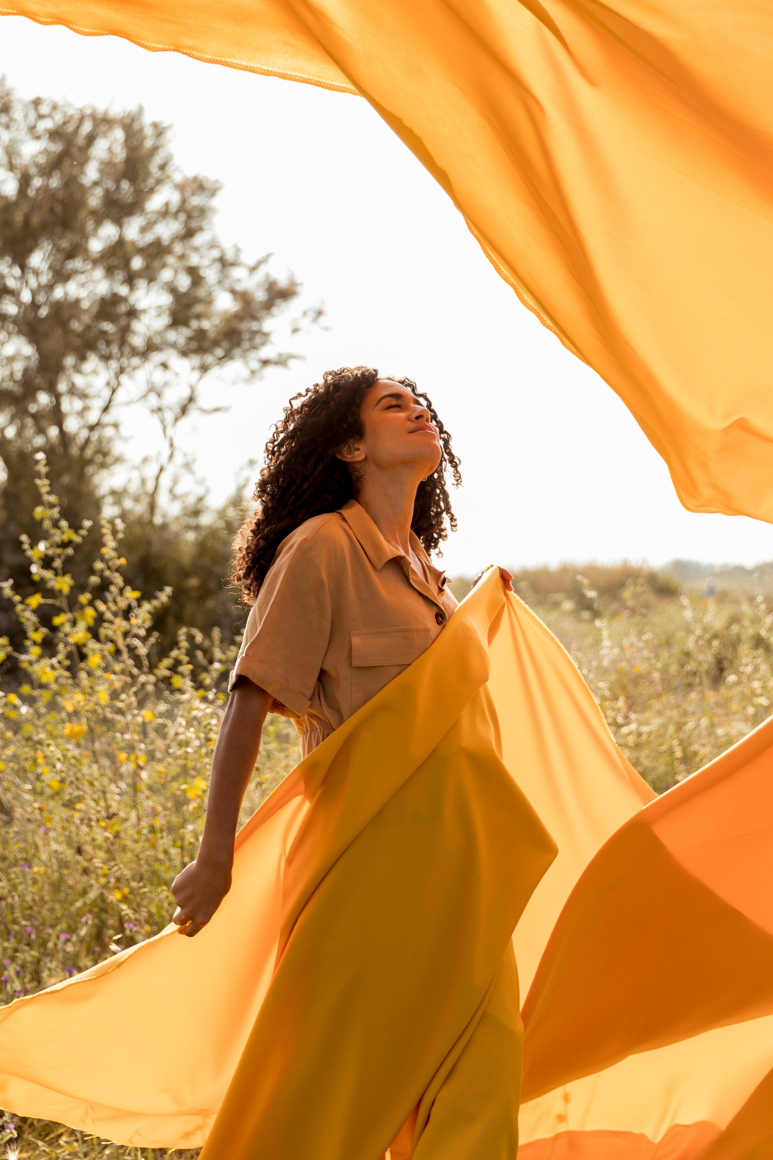 A woman holding flowing yellow fabric outdoors in a field during sunset, with trees in the background.