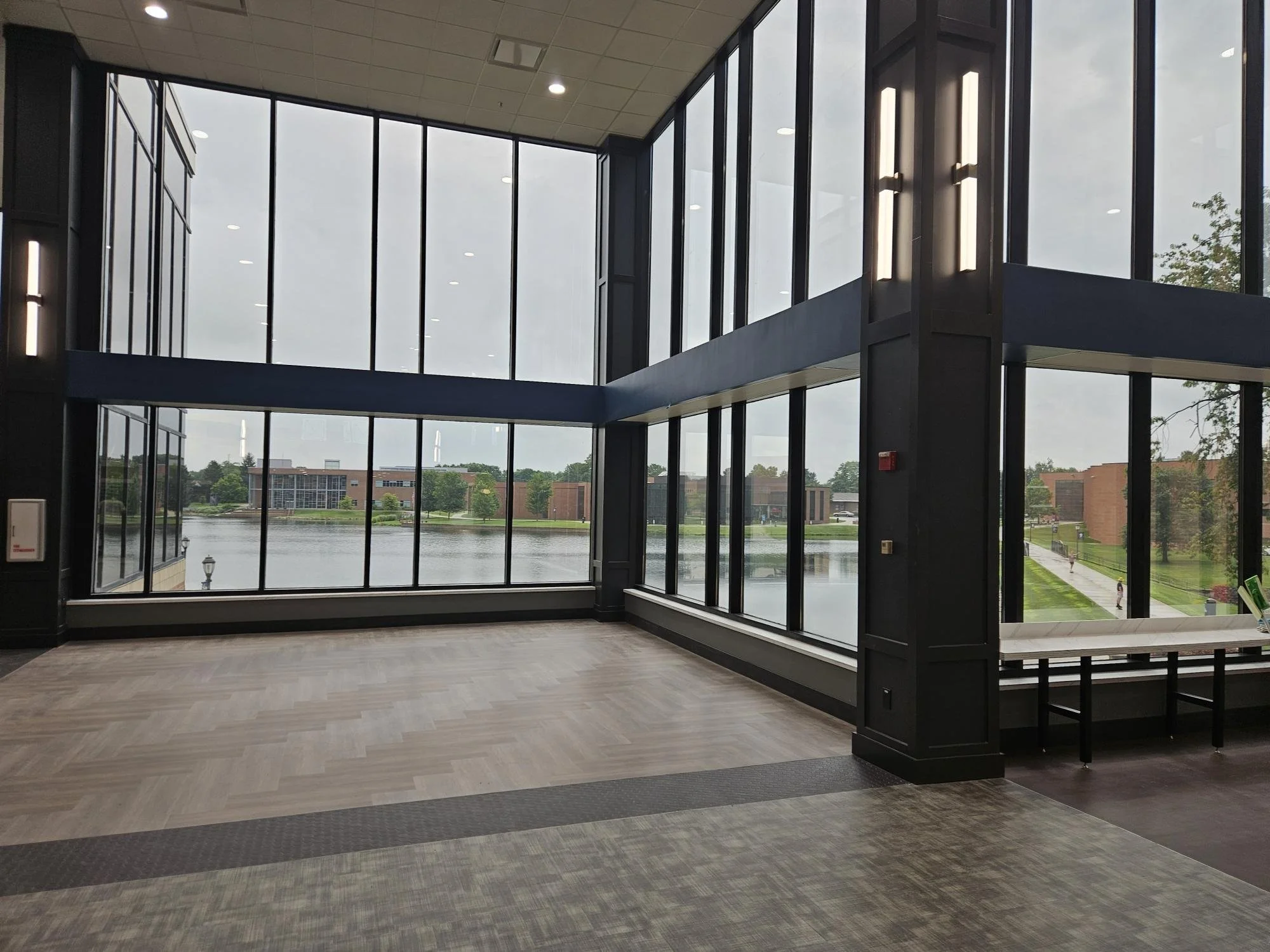 Interior view of a modern building with large glass windows overlooking a lake and campus buildings outside.