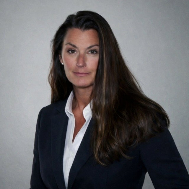 A woman with long brown hair wearing a dark blazer and white shirt, posing against a plain gray background.