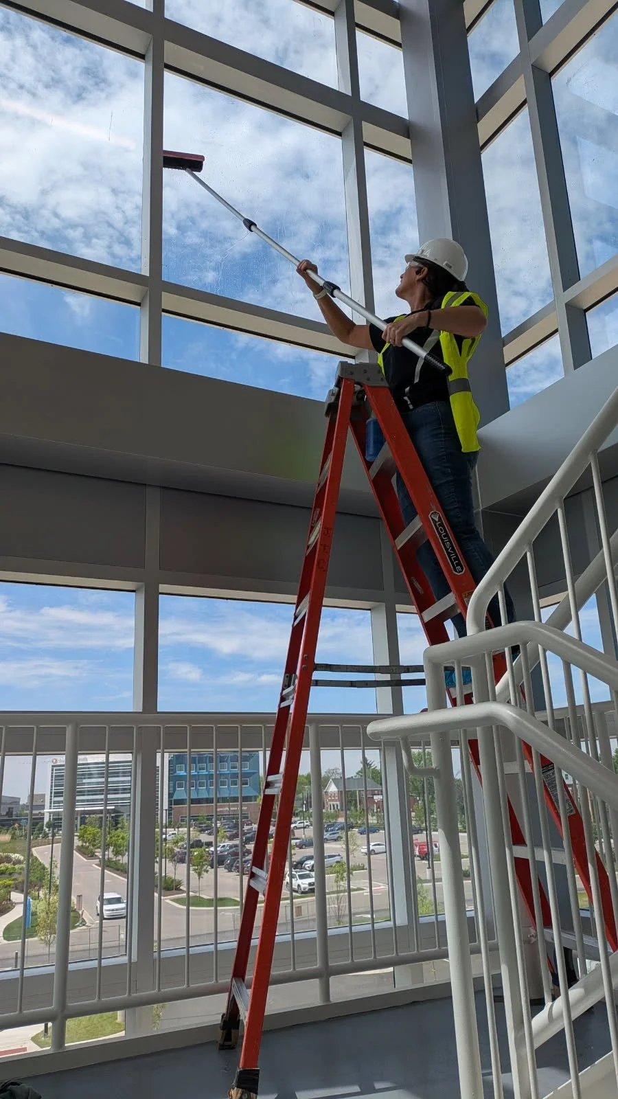 A person standing on a red ladder, cleaning large windows with a long-handled cleaning tool inside a building with a view of a parking lot and modern buildings outside.