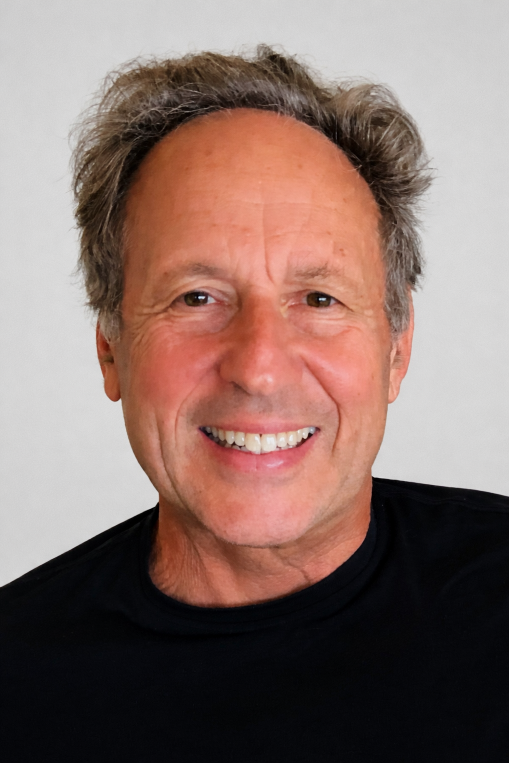 A smiling middle-aged man with wavy, grayish hair, wearing a black shirt, against a plain light background.