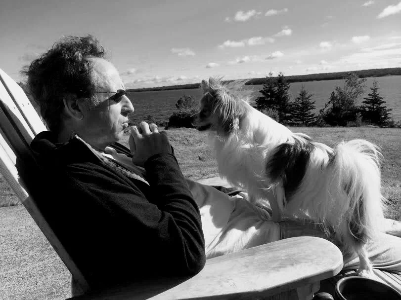 The author, a man, in sunglasses sitting outdoors on a reclining chair, facing a small dog on his lap. They appear to be engaged in a shared moment with a scenic background of water, trees, and a sky with clouds.