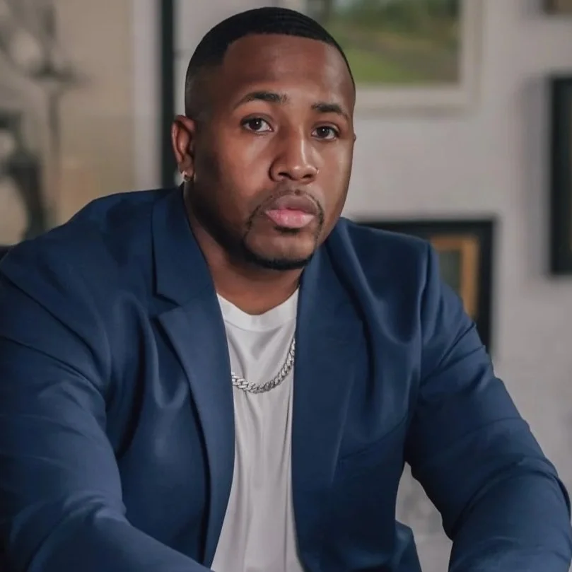 A man with a short haircut and goatee, wearing a navy blazer, white T-shirt, and silver chain, looking directly at the camera in an indoor setting.