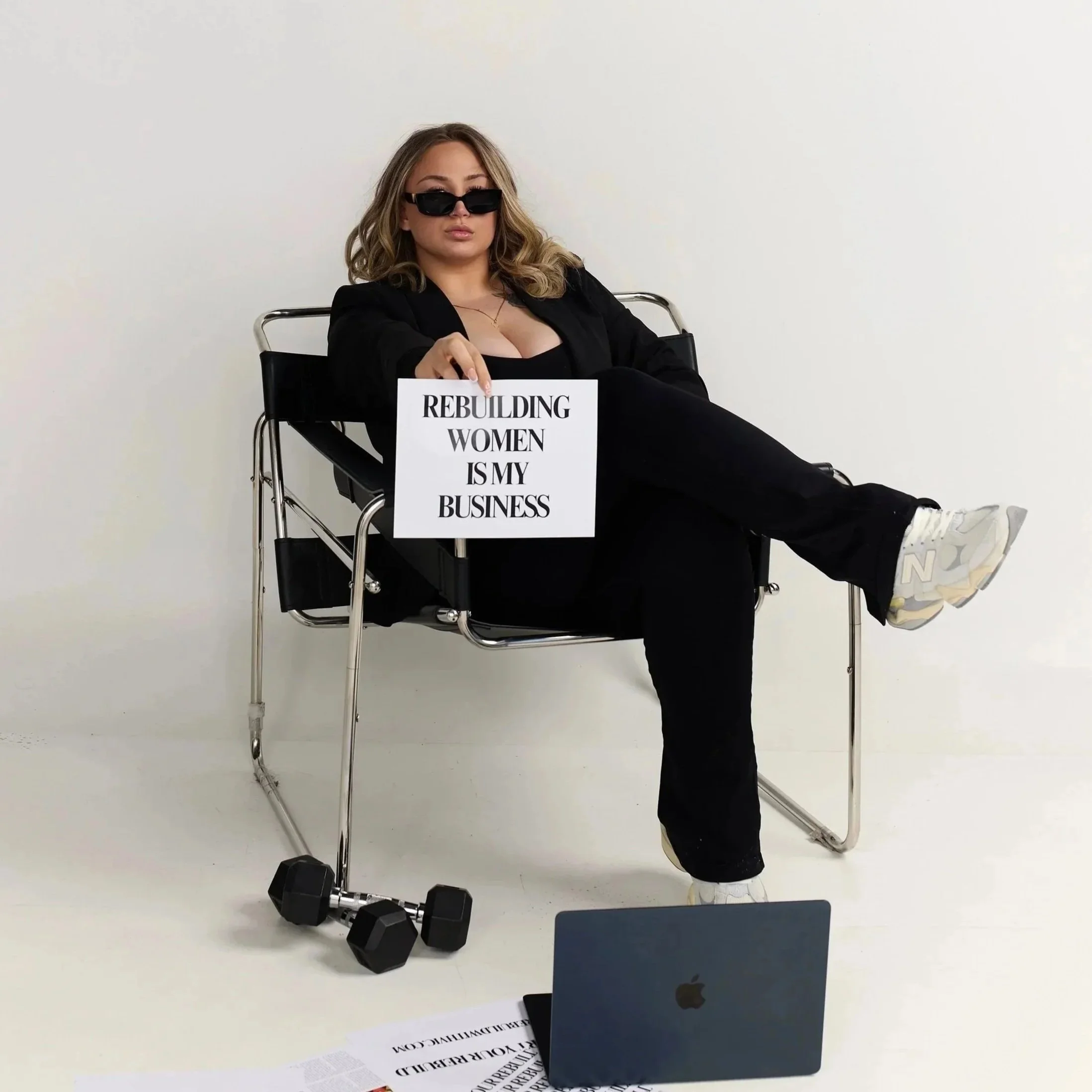 Vic sitting on a modern chair with crossed legs, wearing sunglasses and black clothing, holding a sign that reads, 'Rebuilding women is my business.'