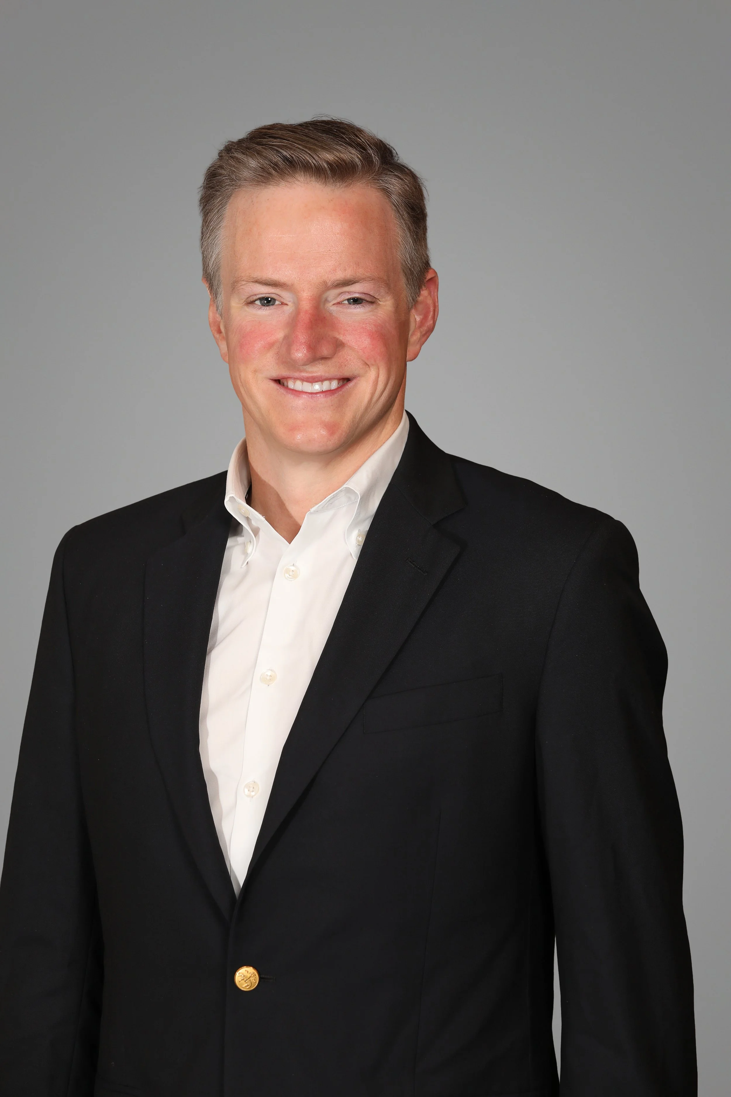 A young man with light skin, short light brown hair, wearing a white dress shirt and black blazer, smiling against a plain gray background.