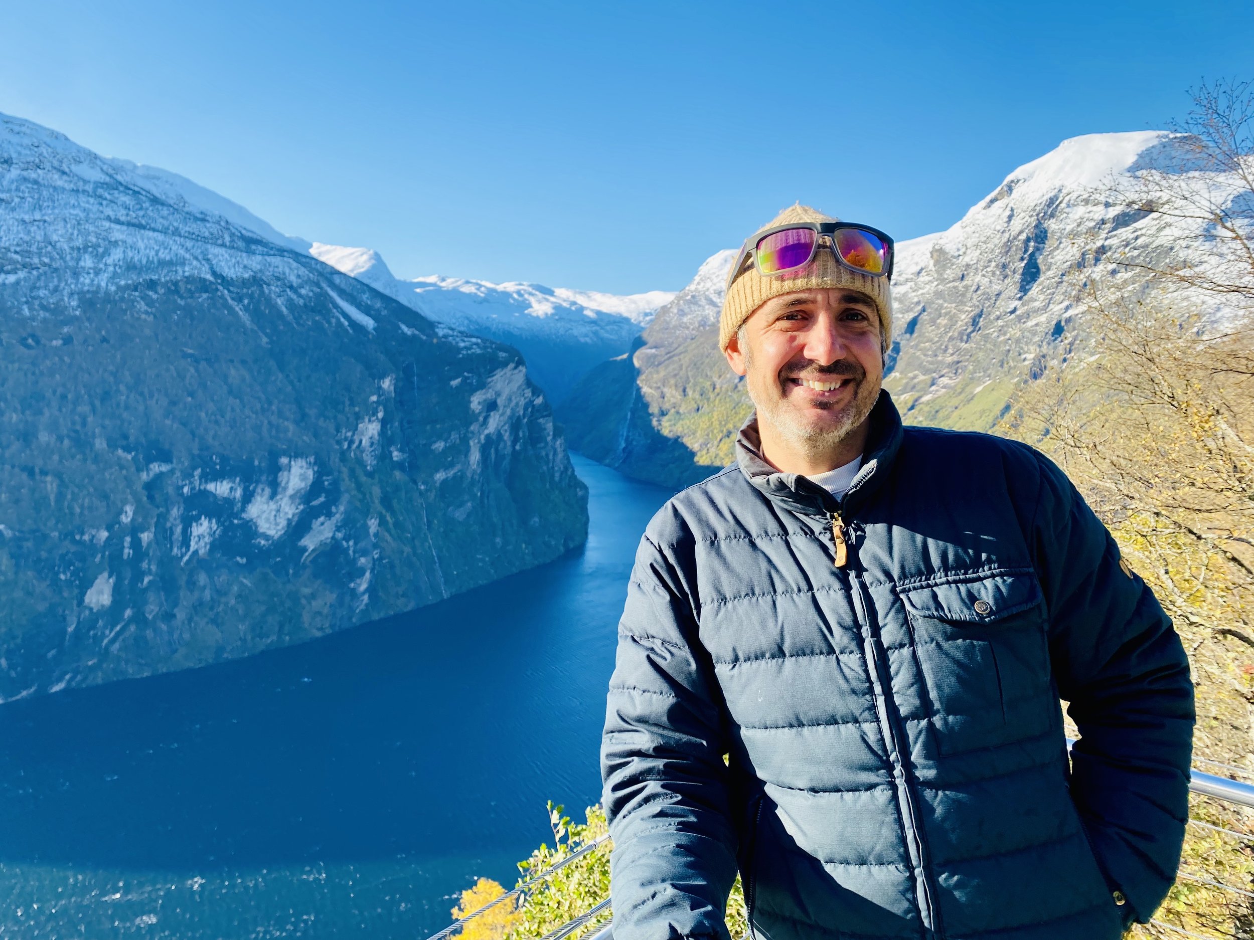 Man smiling in front of a fjord with snow-capped mountains, during a sunny day.