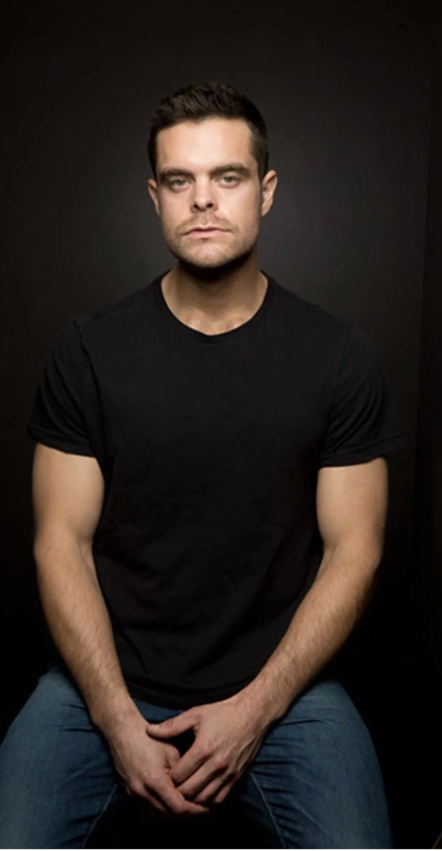 Portrait of a young man with short dark hair, wearing a black t-shirt and sitting against a dark background.